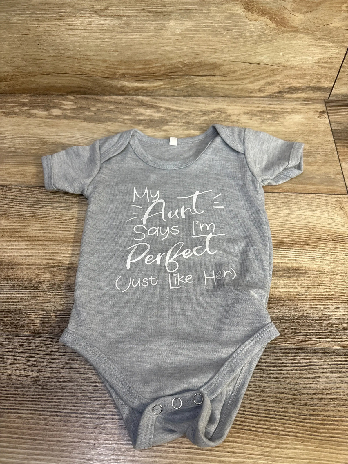 Top-down view of grey baby bodysuit (6 months) showing white script ‘My Aunt Says I’m Perfect (Just Like Her)’ on wood background