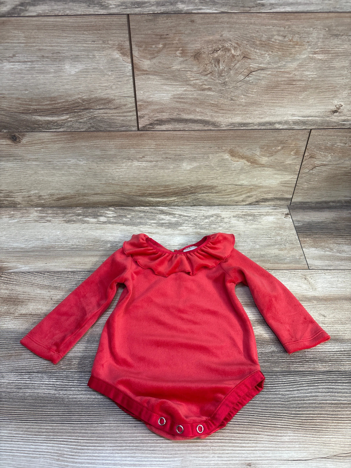 Flat lay of red velour long-sleeve baby bodysuit showing ruffled collar and three snap closures on a wood floor background