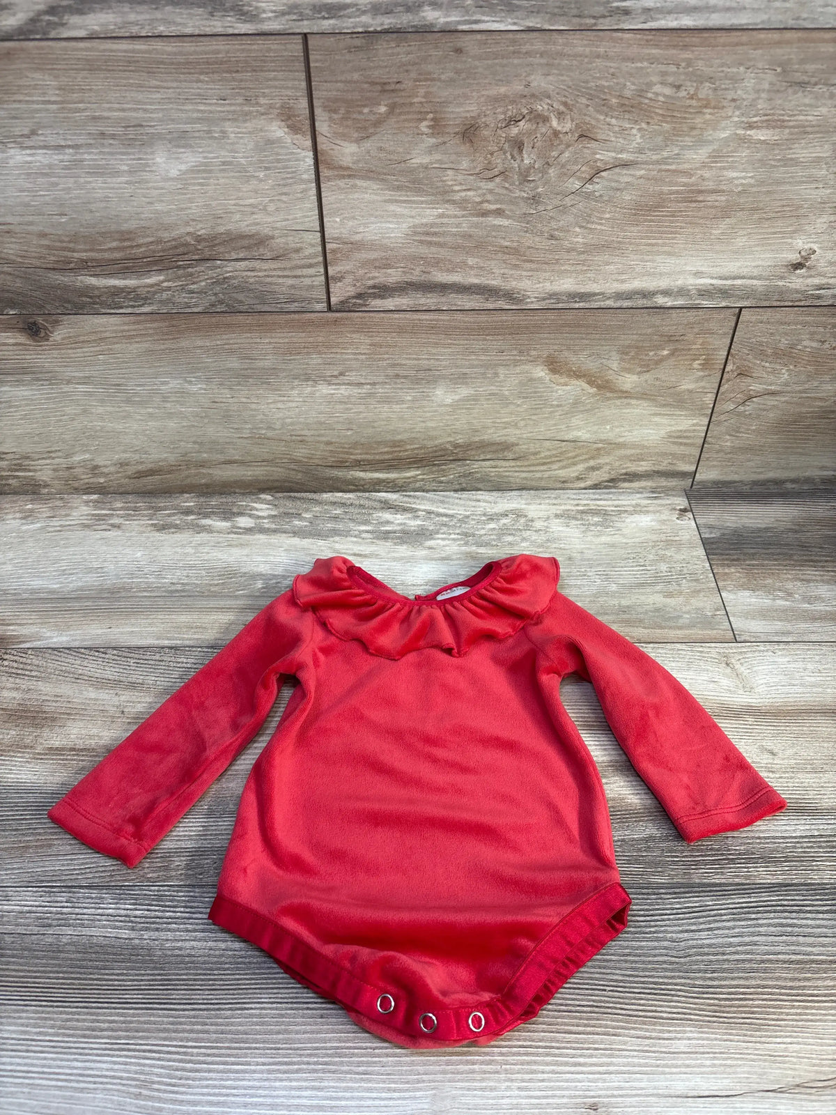 Flat lay of red velour long-sleeve baby bodysuit showing ruffled collar and three snap closures on a wood floor background