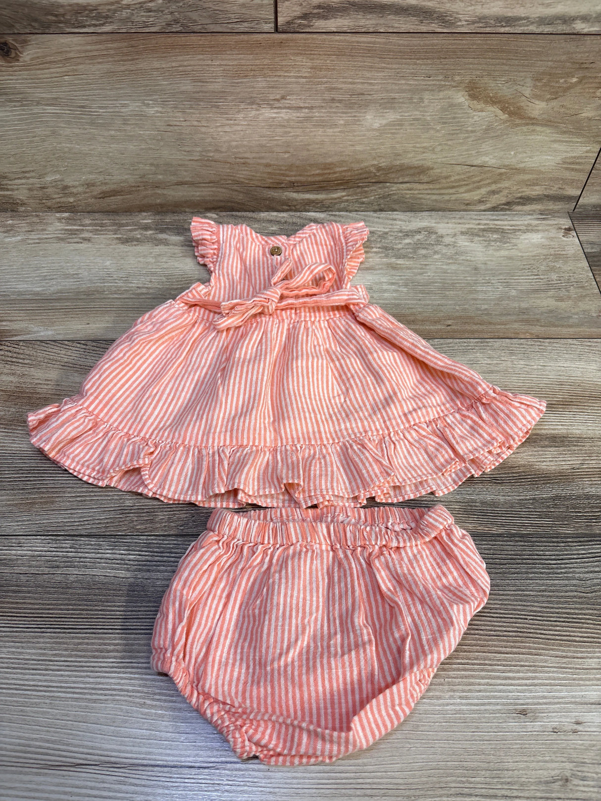 Flat lay of coral and white striped sleeveless baby dress with matching bloomers showcasing ruffle hem, waist tie, and elastic waist on wood background