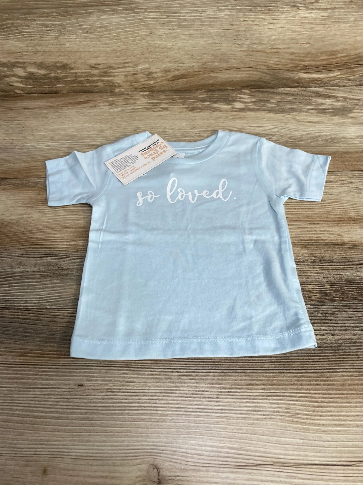 Front view of baby blue Bella Canvas short-sleeve shirt size 6-12 months showing white so loved script on wood background