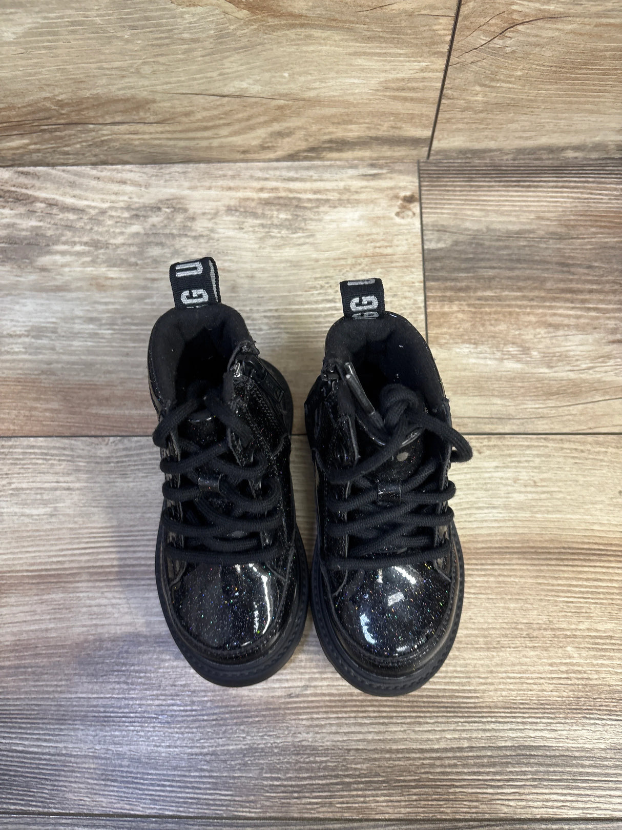 Top-down view of UGG Kids Robley glitter boots showcasing black glitter toe caps and laces on a light wood floor