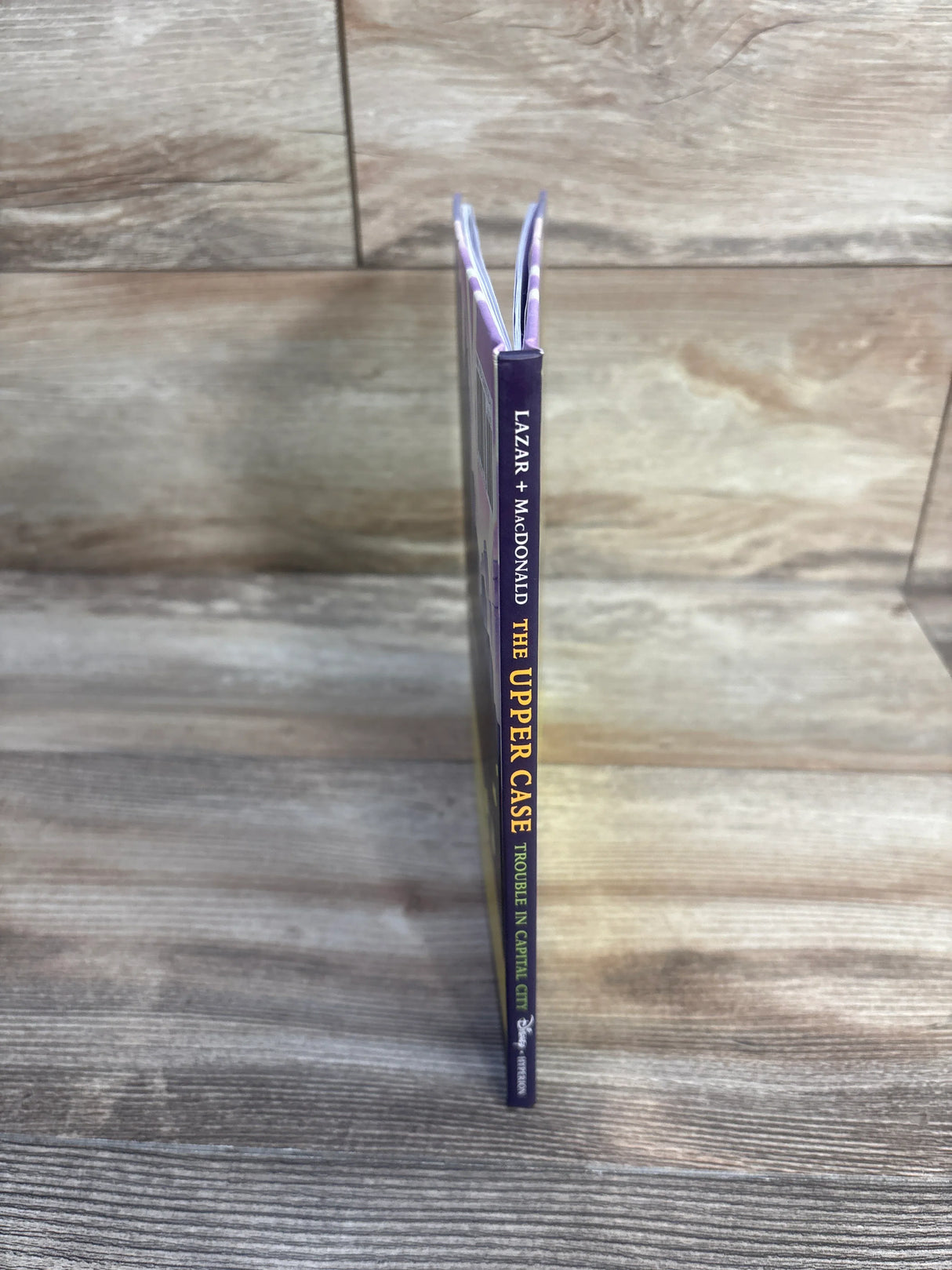 Spine view of The Upper Case: Trouble in Capital City hardcover book showcasing its purple spine against a wood background