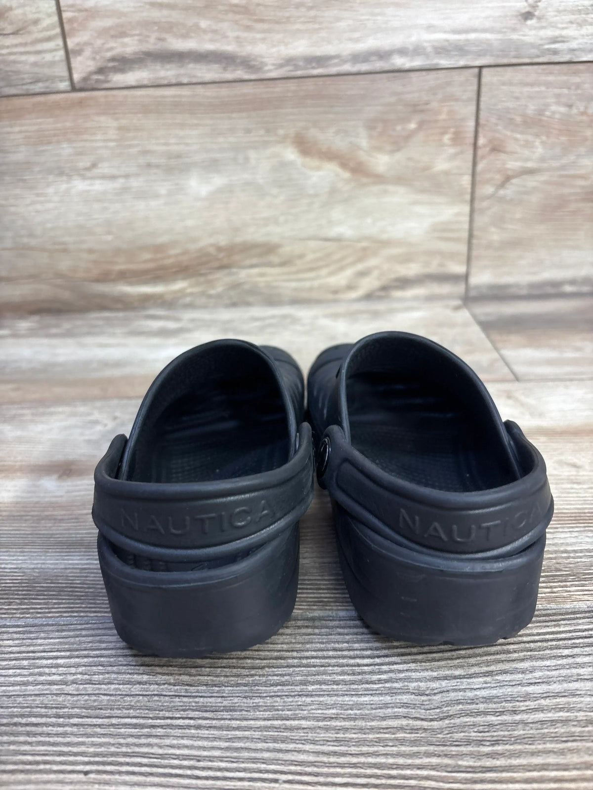 Rear view of Nautica River Edge clogs showing embossed ‘NAUTICA’ heel strap and molded black rubber construction on wood-look floor