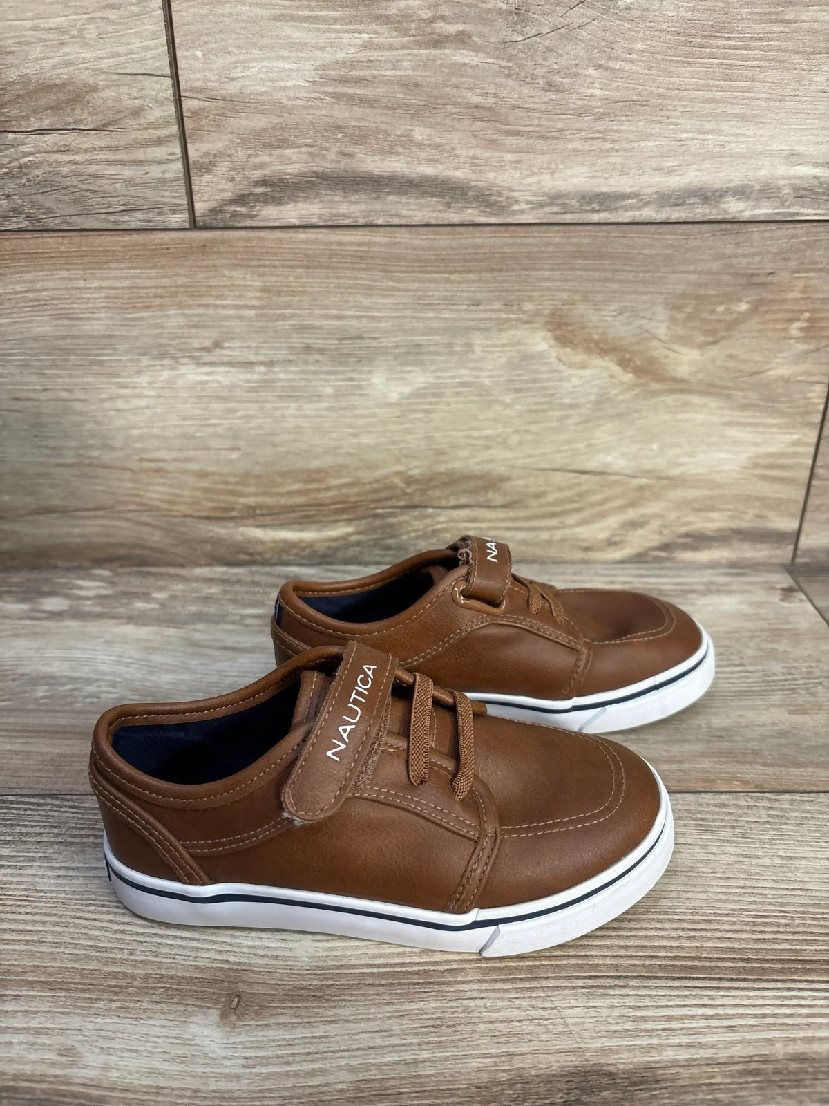 Side view of Nautica Calloway faux leather brown sneakers showcasing lace-up fronts, Nautica straps, and white soles on a wood-look floor