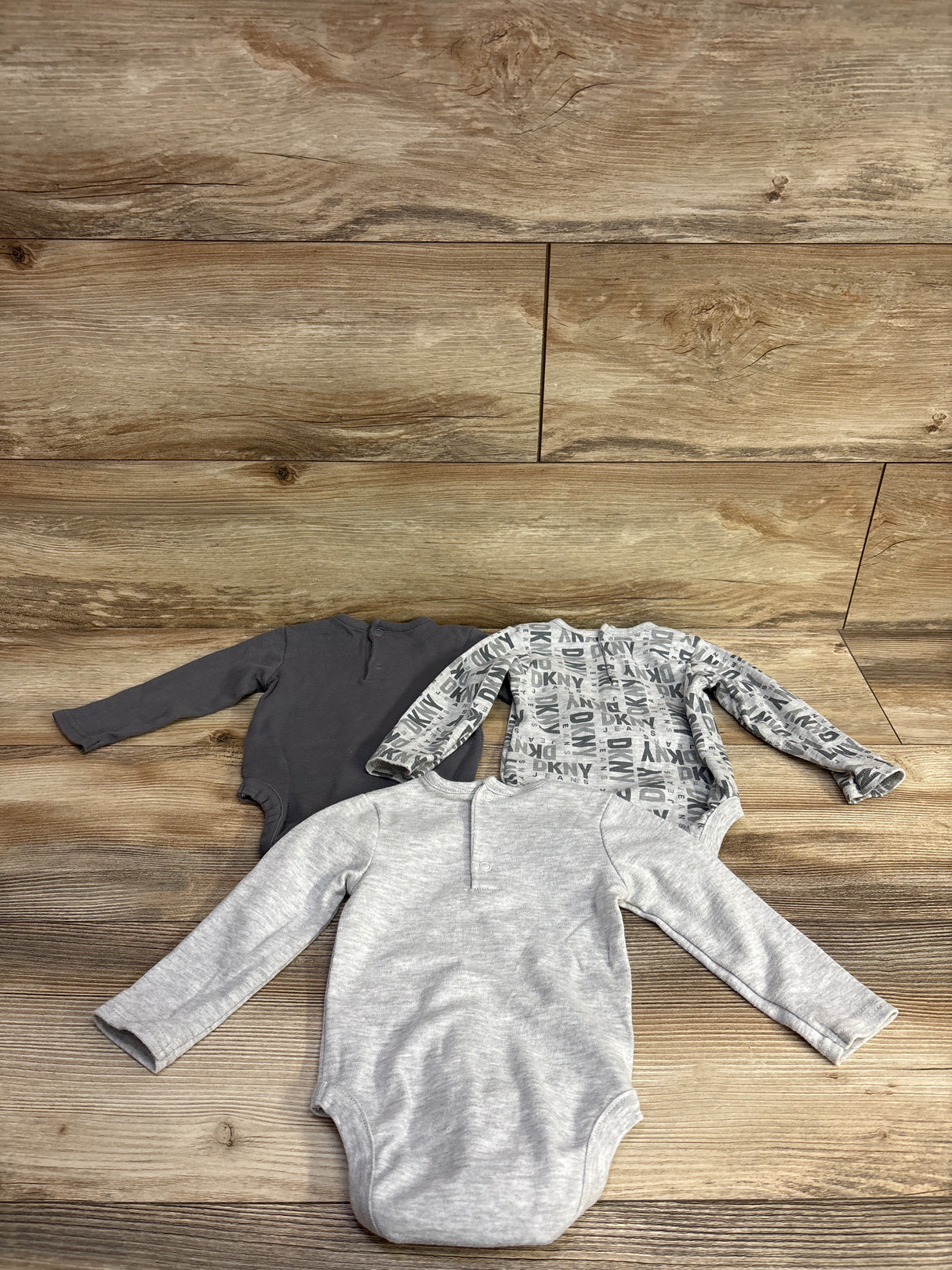 Three long-sleeve baby bodysuits in gray, patterned gray with DKNY logo, and dark gray on wooden floor