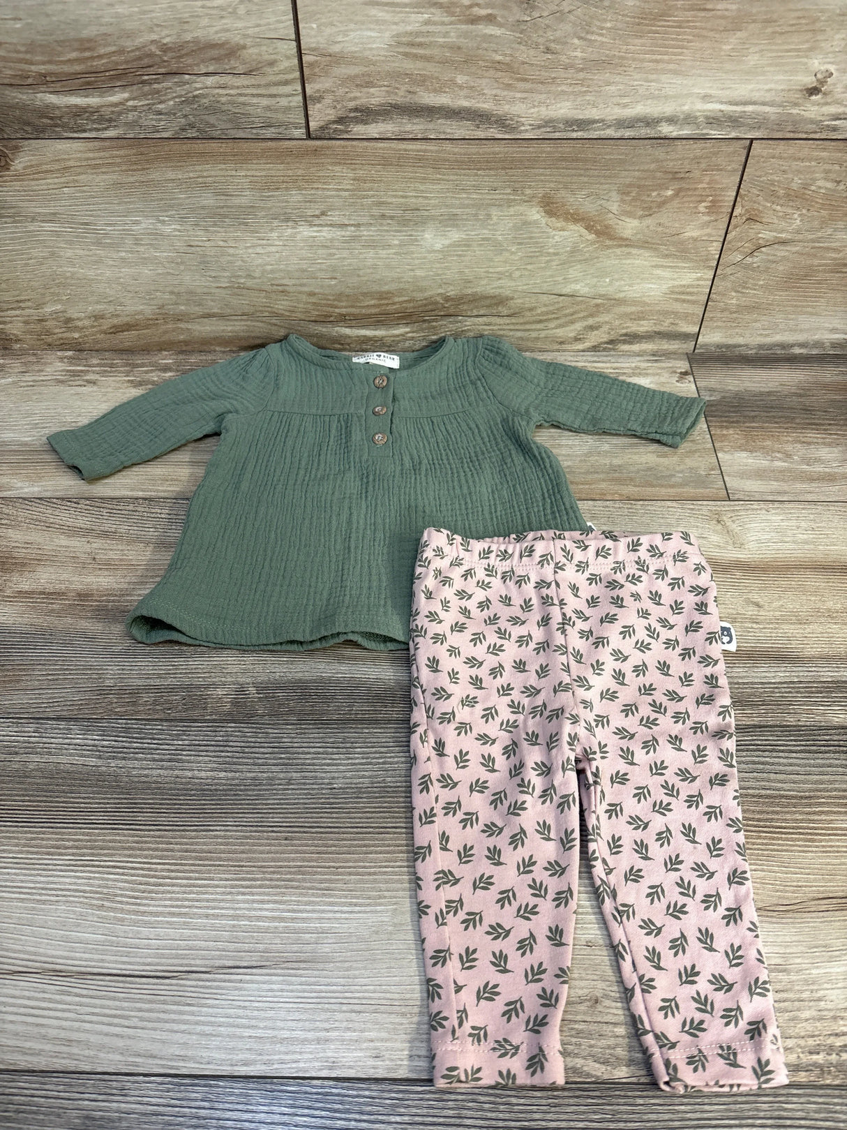 Green long-sleeve baby top with buttons and pink leaf-patterned leggings on wood floor