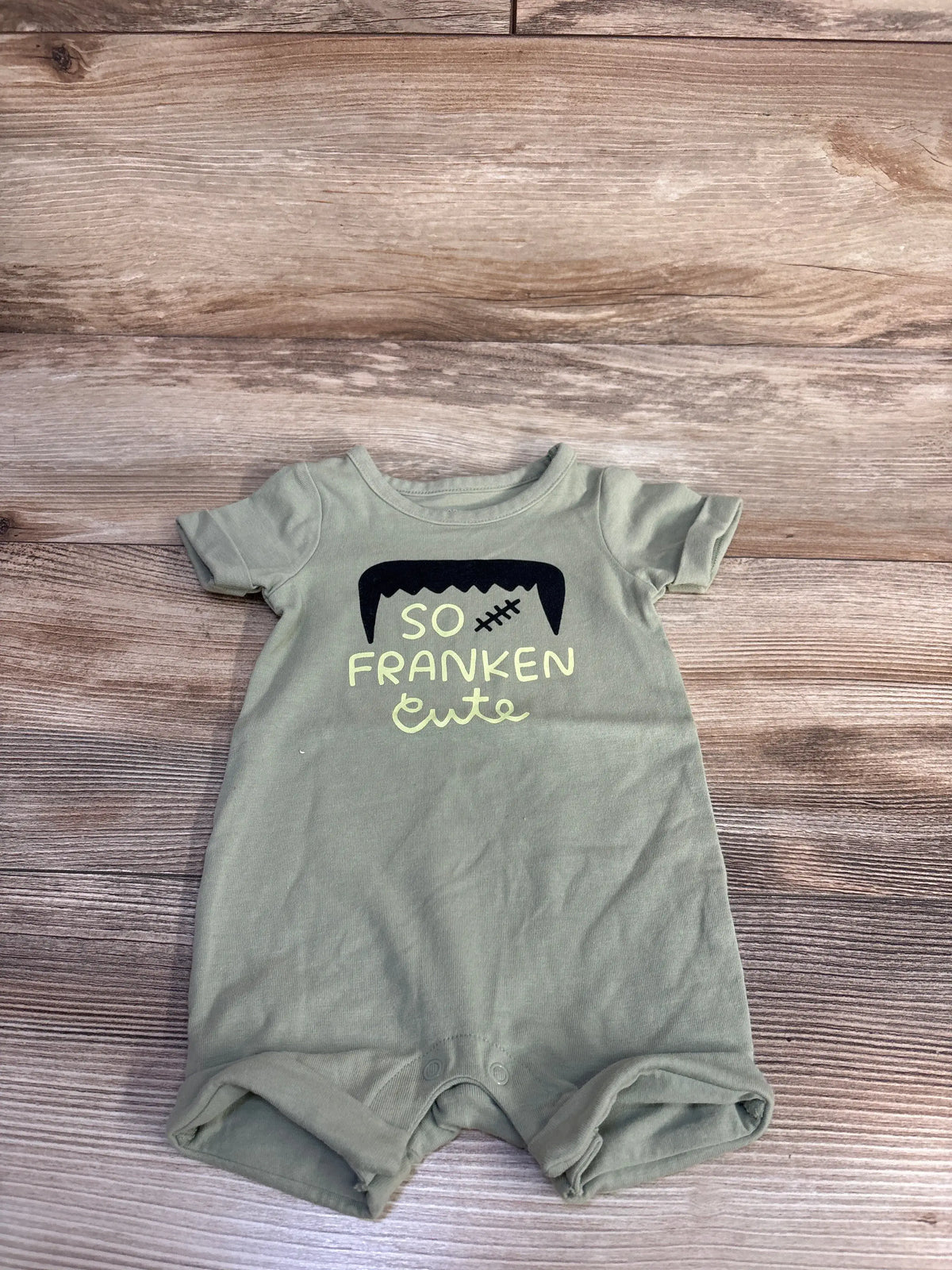 Front view of Cat & Jack green shortie romper, size 0 to 3 months, with So Franken Cute graphic on wood-look background