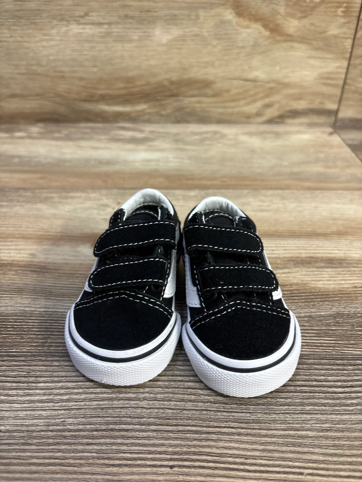 Front view of black Vans Old Skool V sneakers in size 2C showcasing hook-and-loop straps and white soles on a wood floor