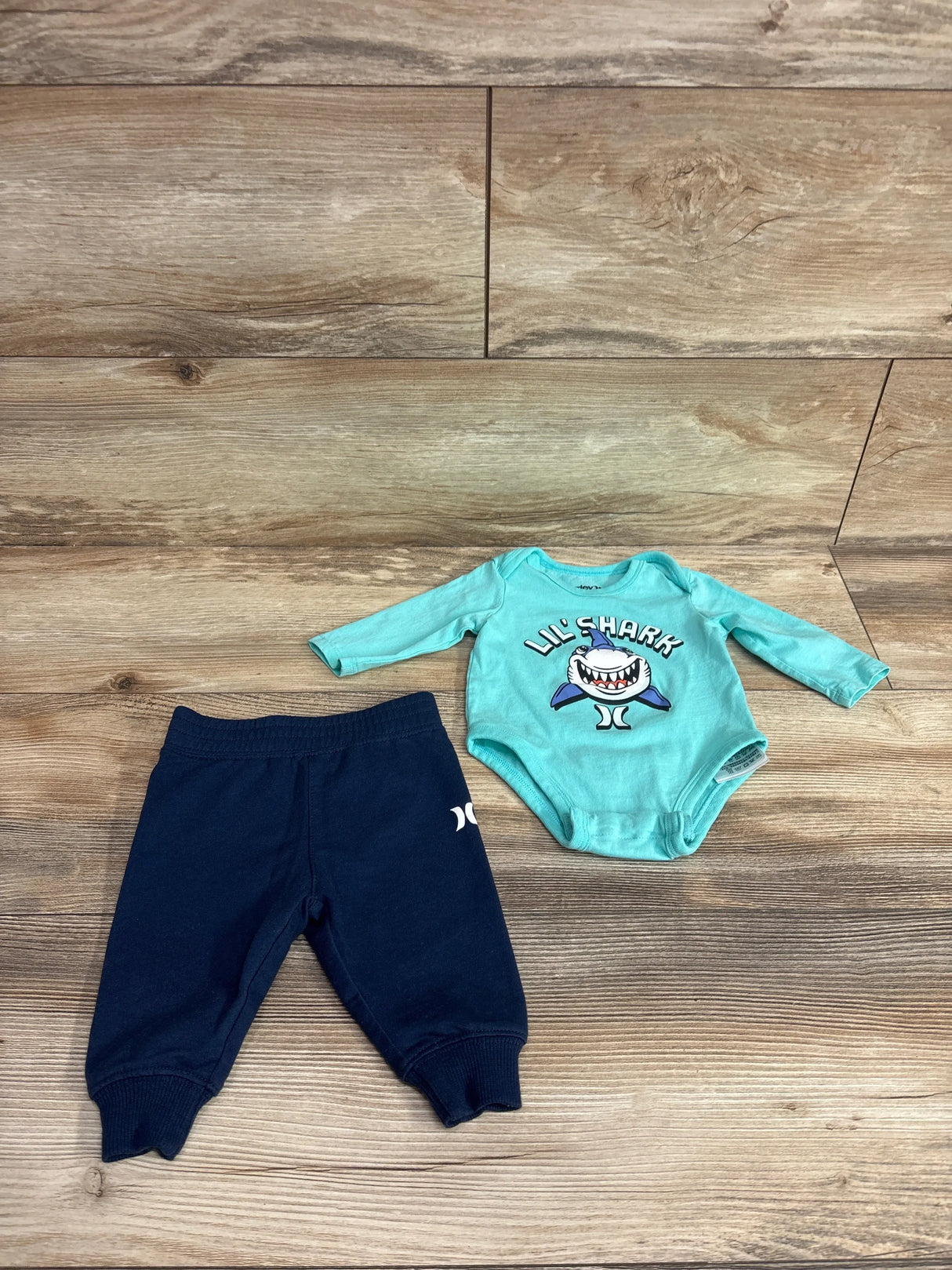 Flat lay of Hurley 2-piece baby outfit showing mint green Lil Shark long-sleeve bodysuit and navy pants on wood background