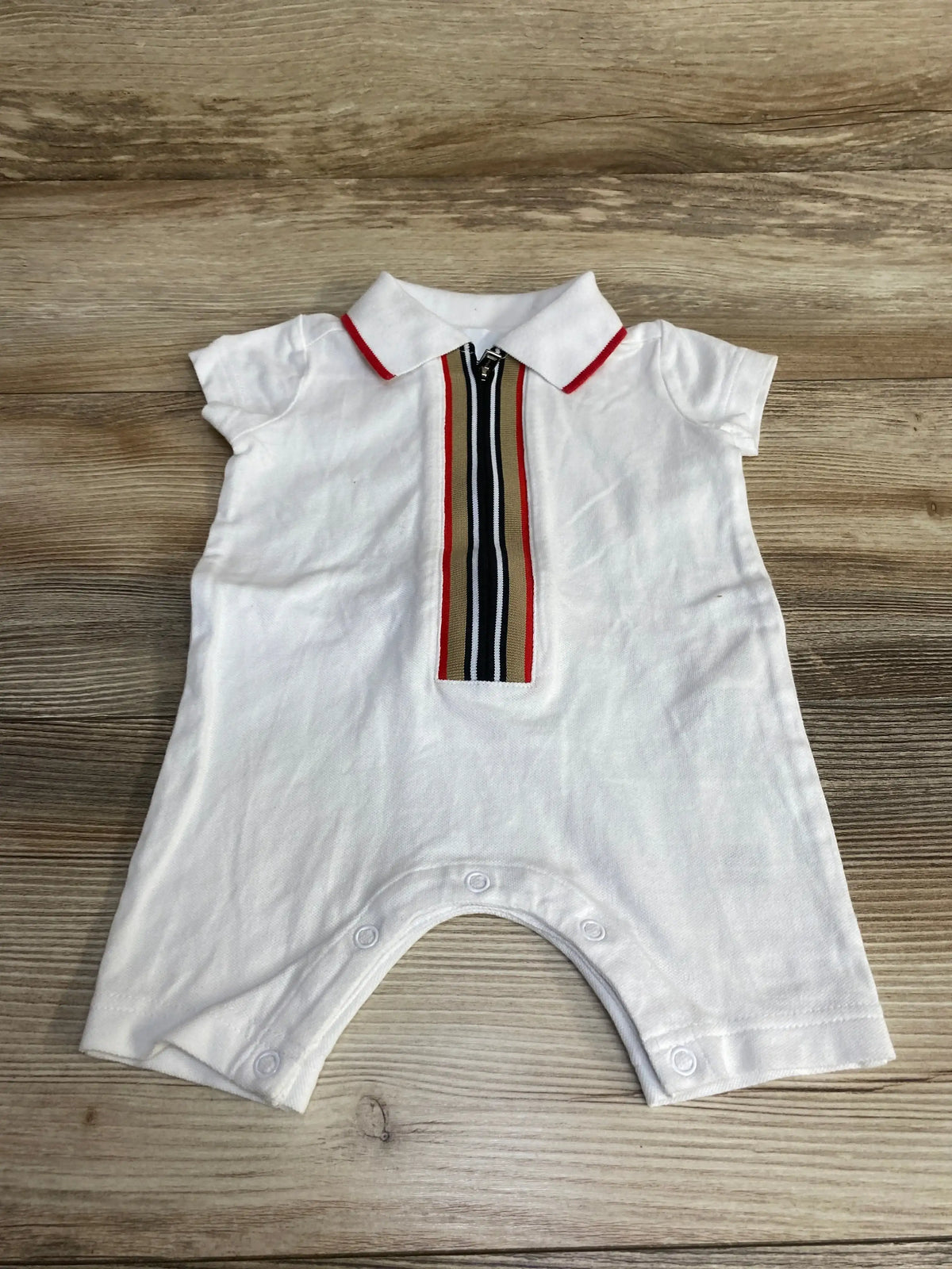 Flat lay of white Burberry polo romper with red-trim collar, signature stripe placket and snap crotch on wood background