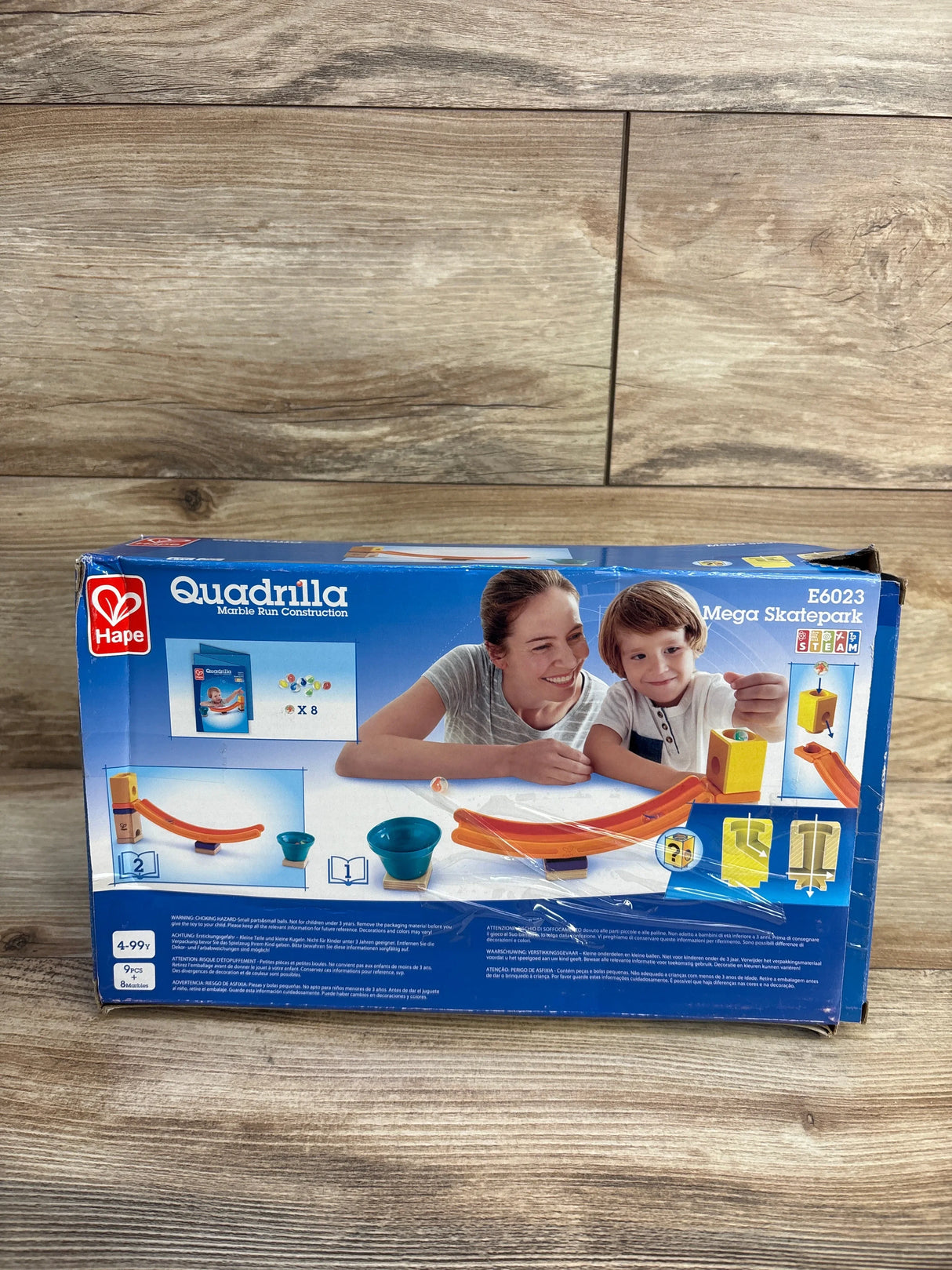 Front view of Hape Quadrilla Mega Skatepark wooden marble-run playset box showing illustrated parent and child, orange curved tracks, and blue bowls on wood floor background