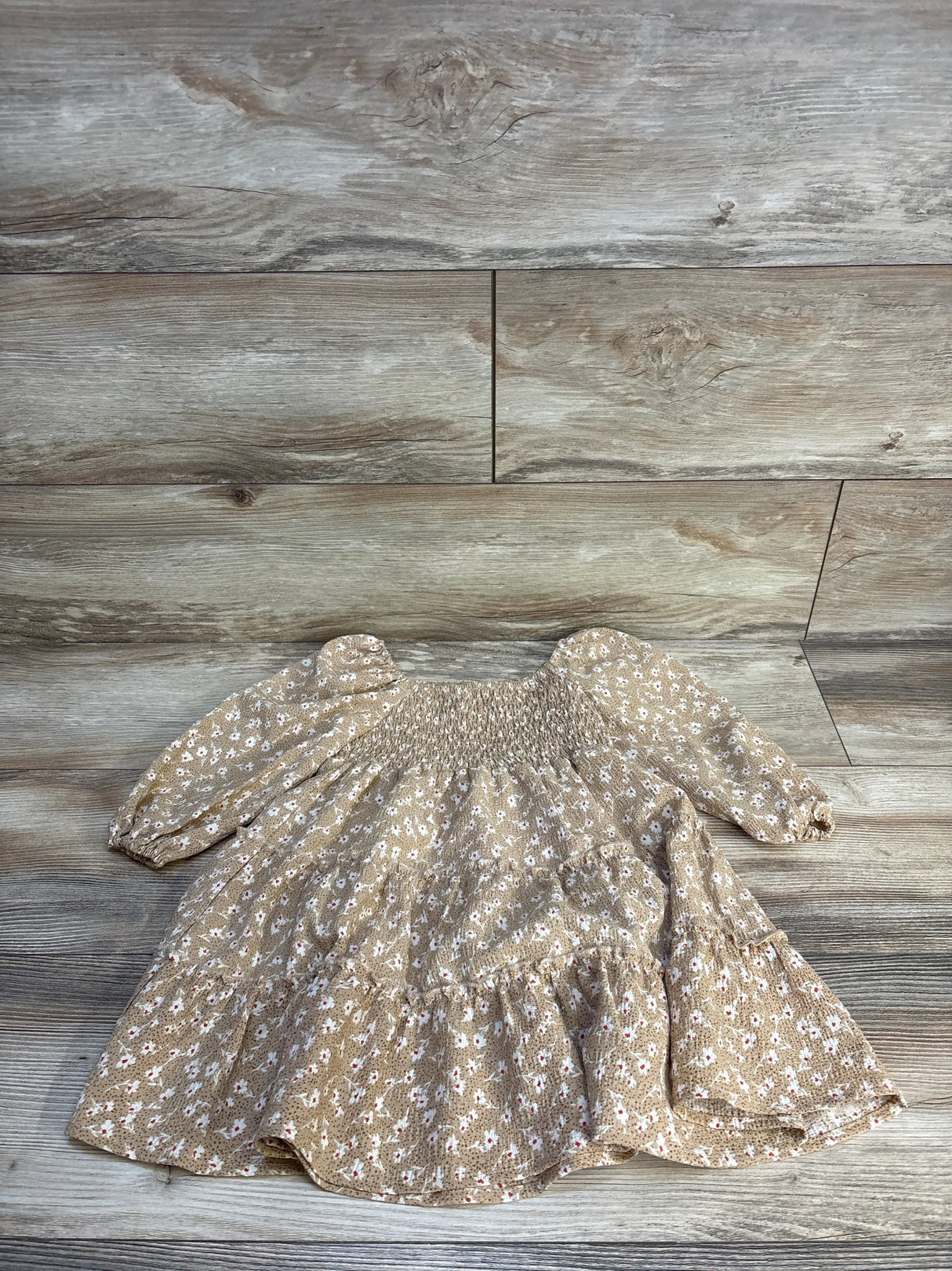 Tan floral baby dress with smocked bodice and long sleeves, laid on wood floor