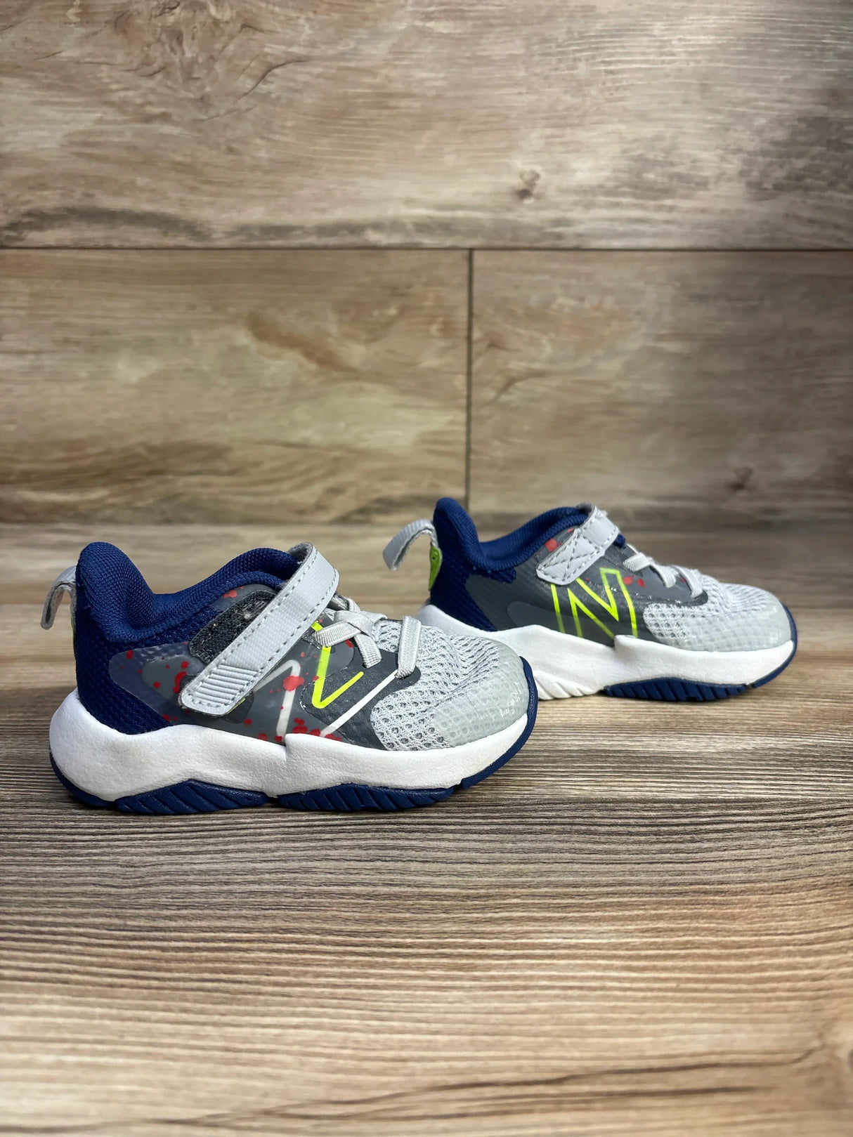 Side view of New Balance Rave Run v2 kids sneakers in white and blue with mesh upper, Velcro strap and neon logo on wood floor