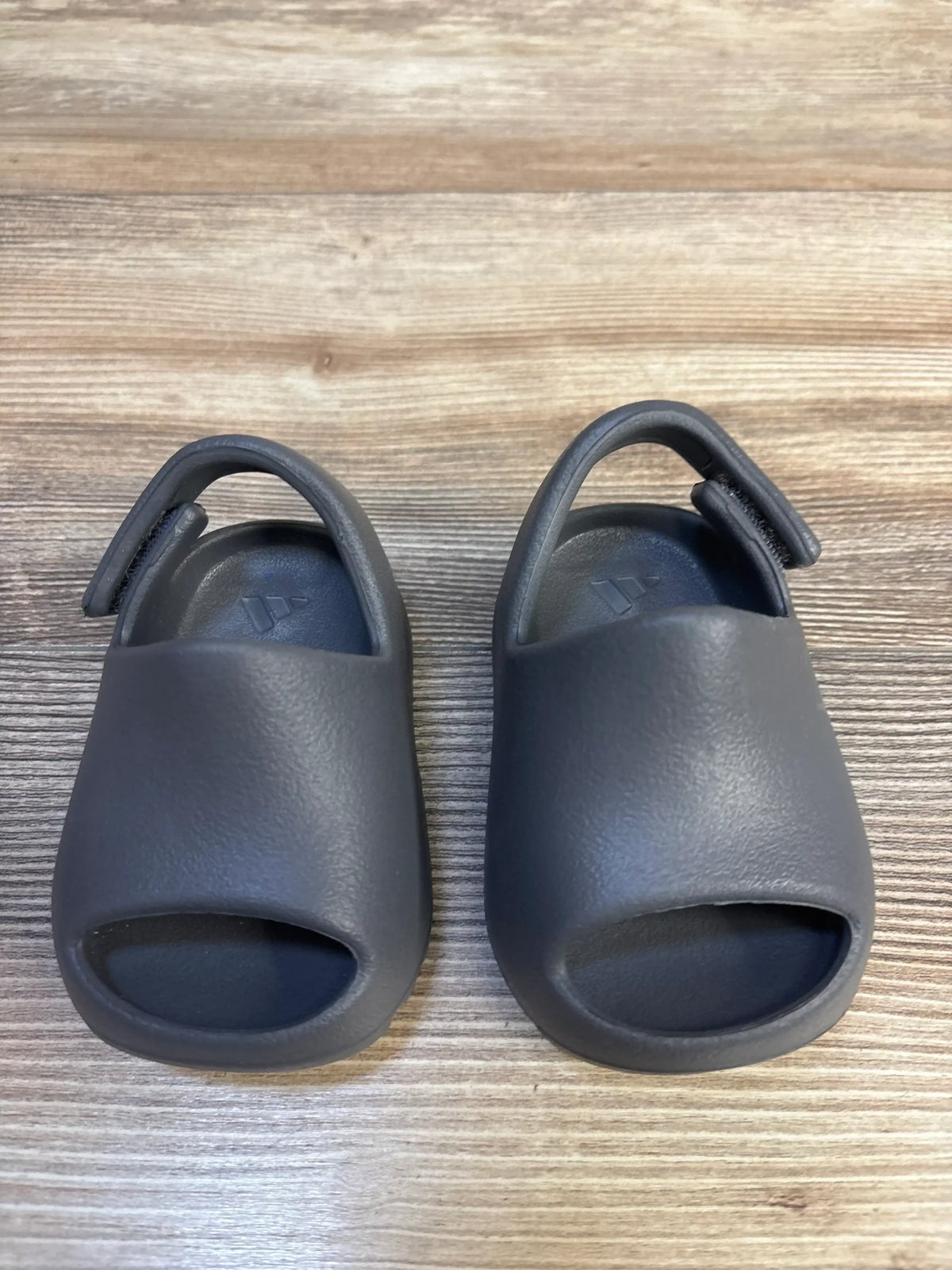 Top view of Adidas Yeezy Slides in Granit, toddler size 3C, showing open toe and adjustable heel straps on wood surface