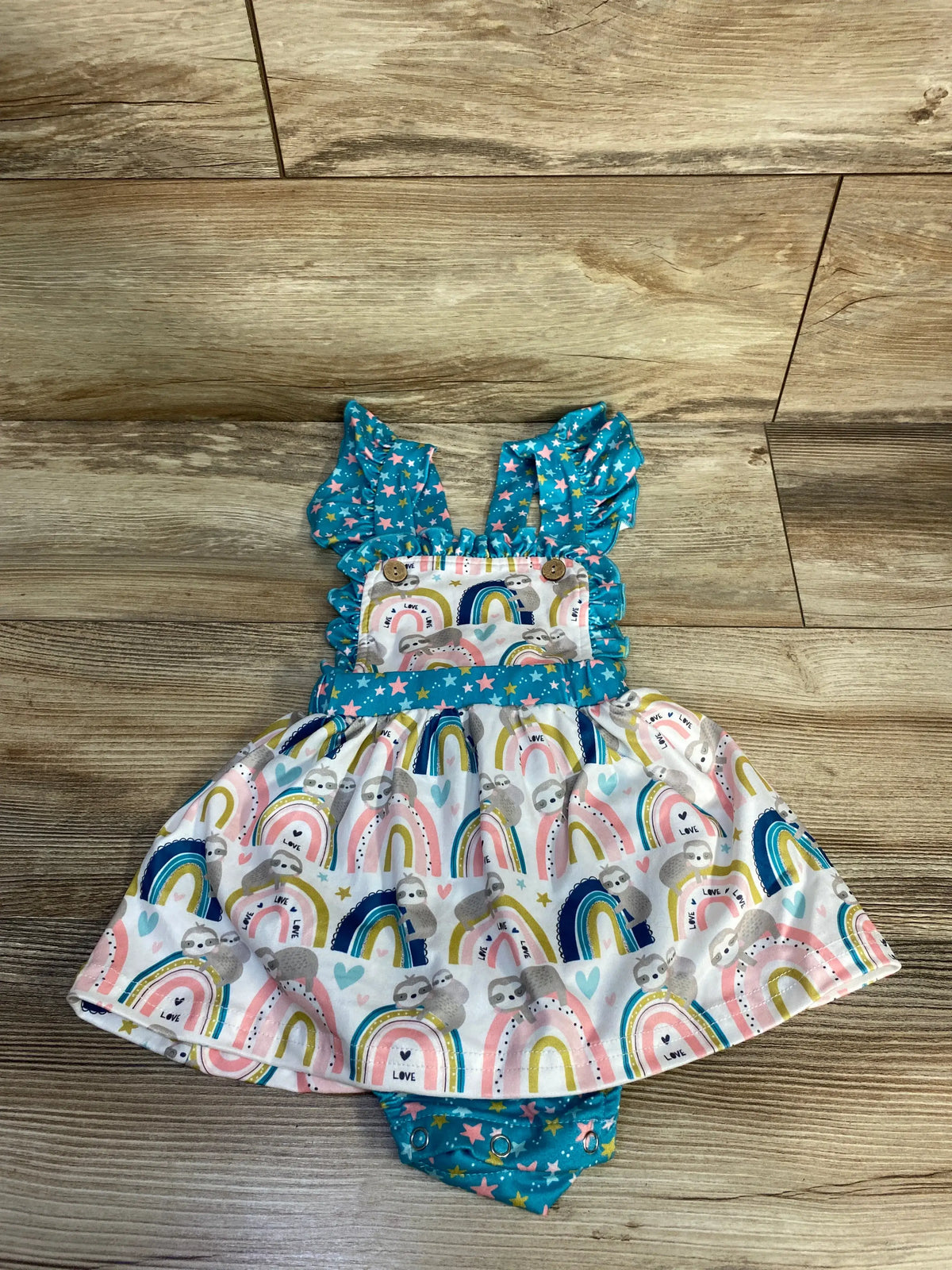 Front view of Pete + Lucy sloth bodysuit dress with rainbow sloth print, blue straps, and snap closure on wood flooring