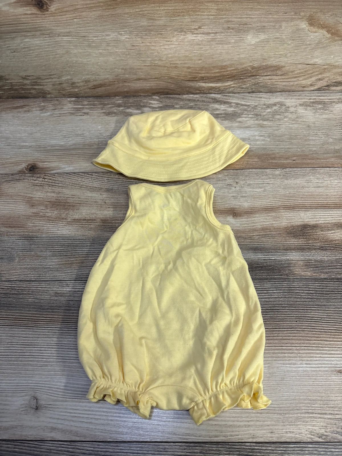 Top-down view of yellow two-piece romper and hat set showcasing a sleeveless romper and matching hat on a wood-grain background