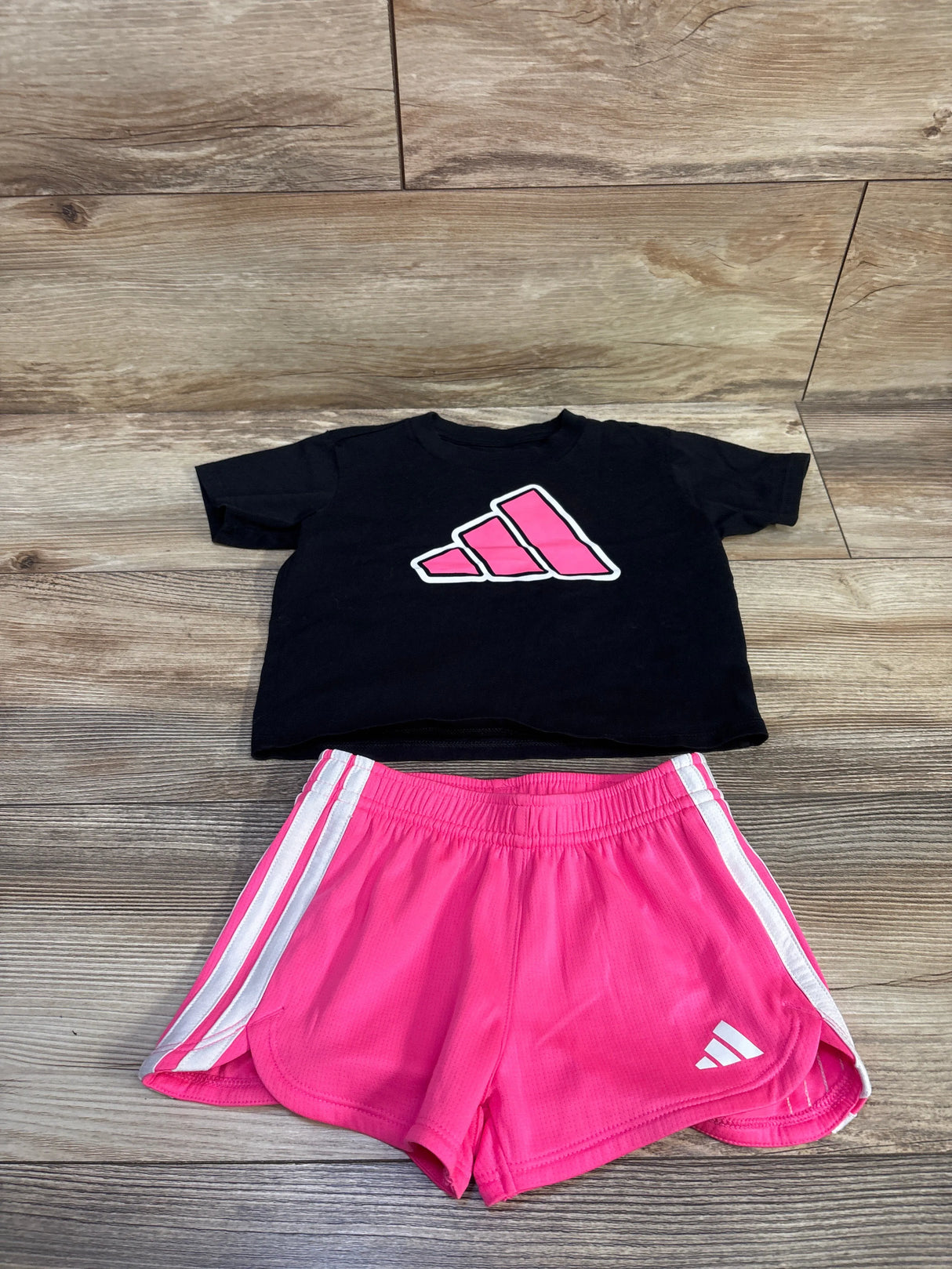 Flat lay of Adidas toddler two-piece set: black cropped tee with pink logo and pink shorts with white side stripes on wood