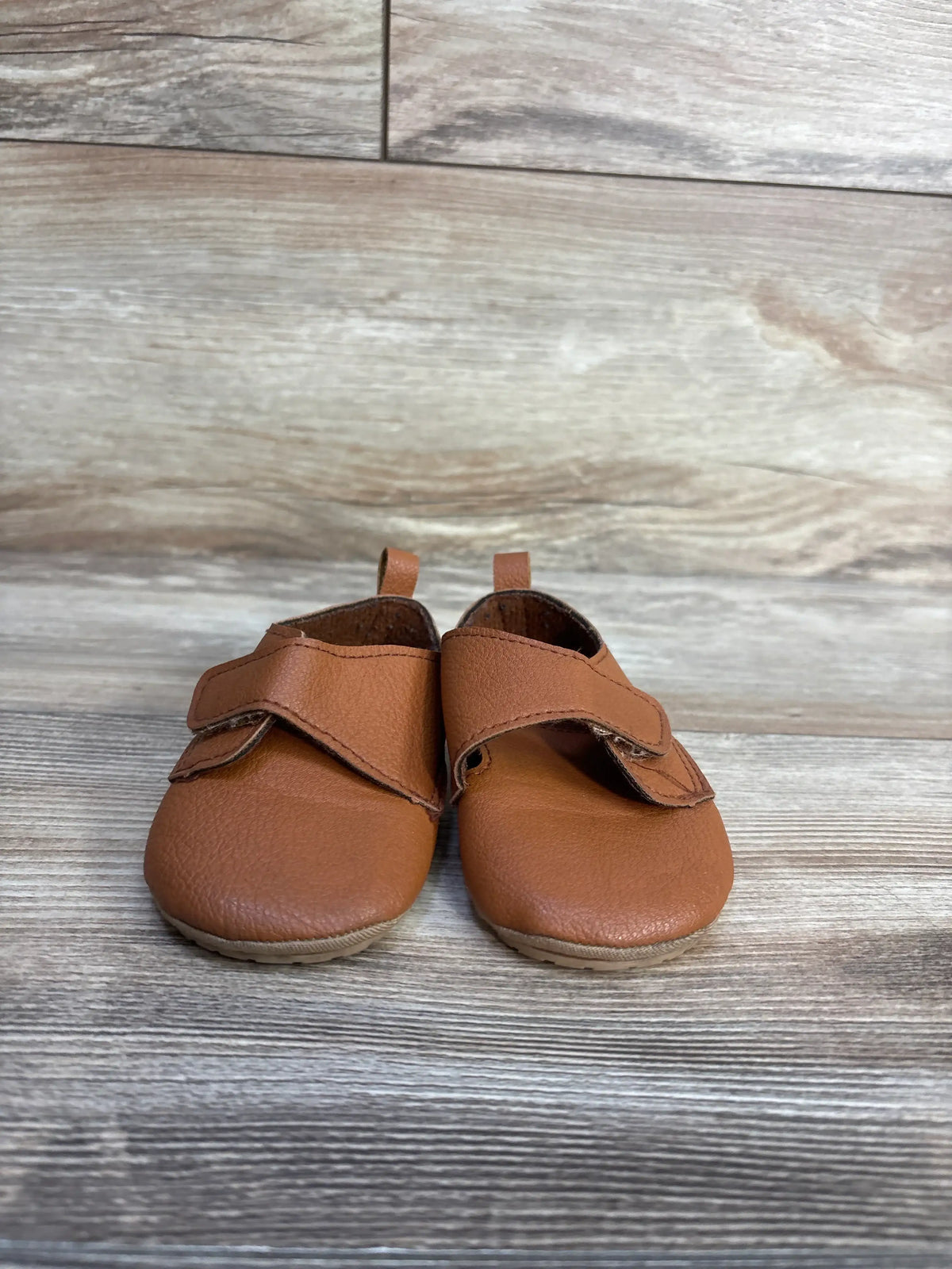 Front view of brown faux leather first walking soft sole shoes showcasing hook and loop closure and pull tabs on wood floor