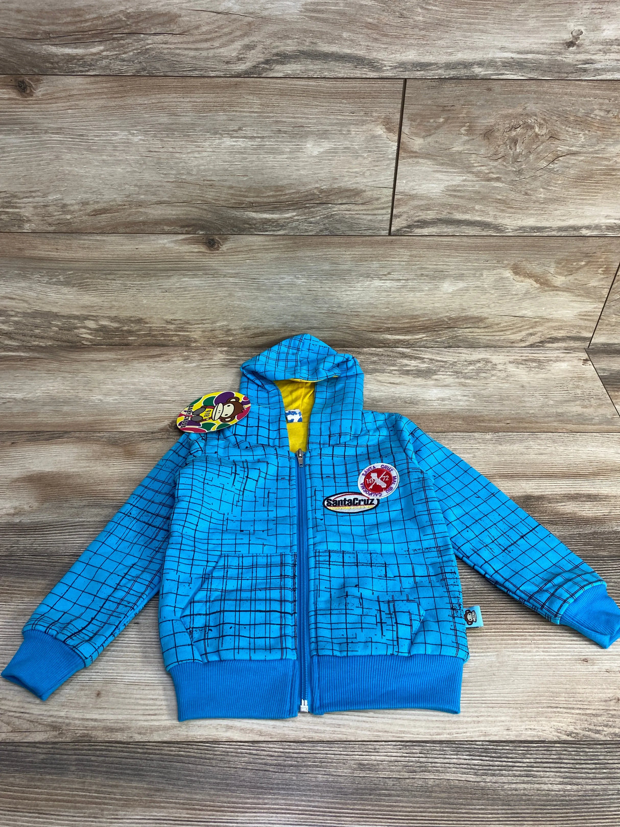 Blue children's zip-up hoodie with grid pattern, Santa Cruz patches, and yellow hood lining on wooden floor
