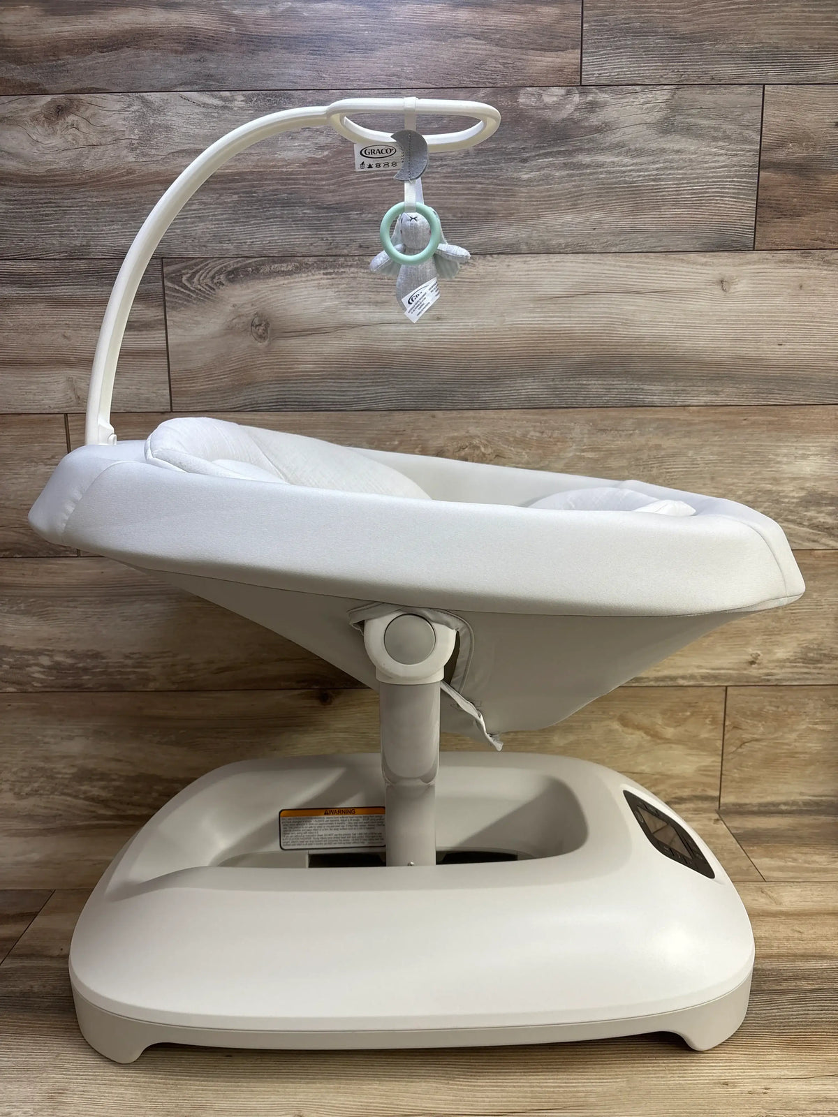 Front three-quarter view of Graco Smart Sense Soothing Baby Swing in white with padded seat, toy bar, and wide base on a wood floor