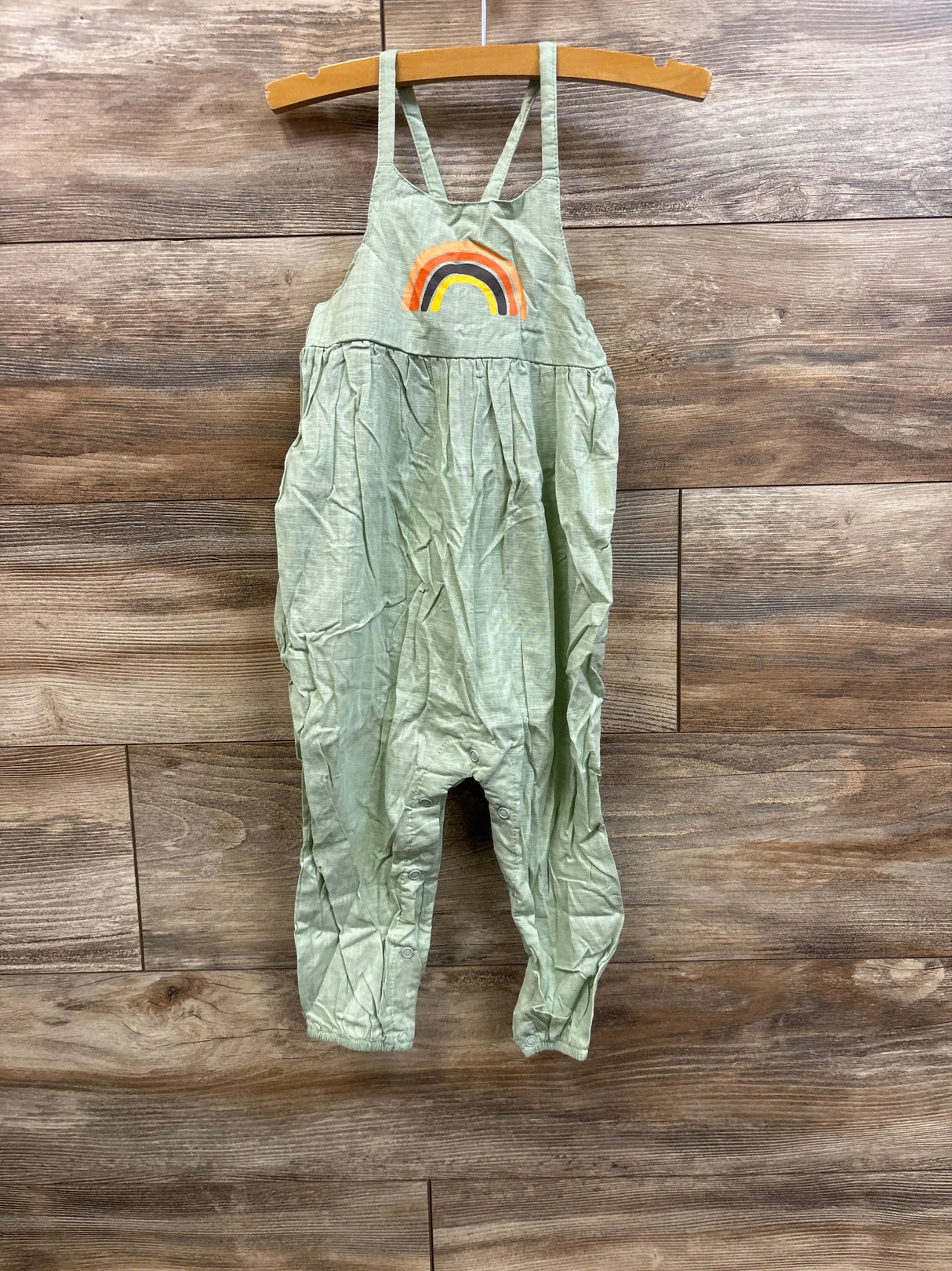 Front view of green toddler jumpsuit with rainbow applique and snap closures at legs on wooden backdrop