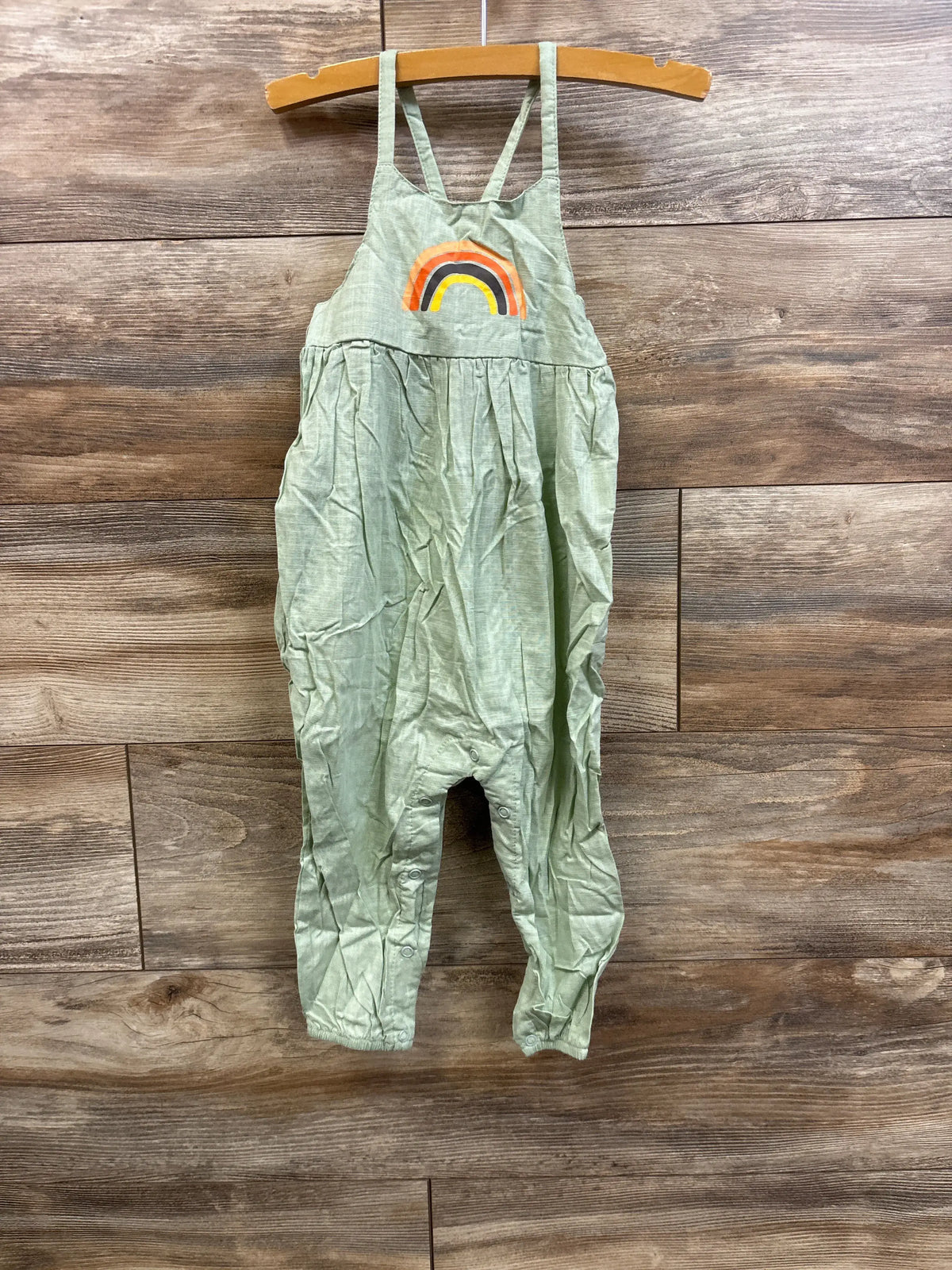 Front view of green toddler jumpsuit with rainbow applique and snap closures at legs on wooden backdrop