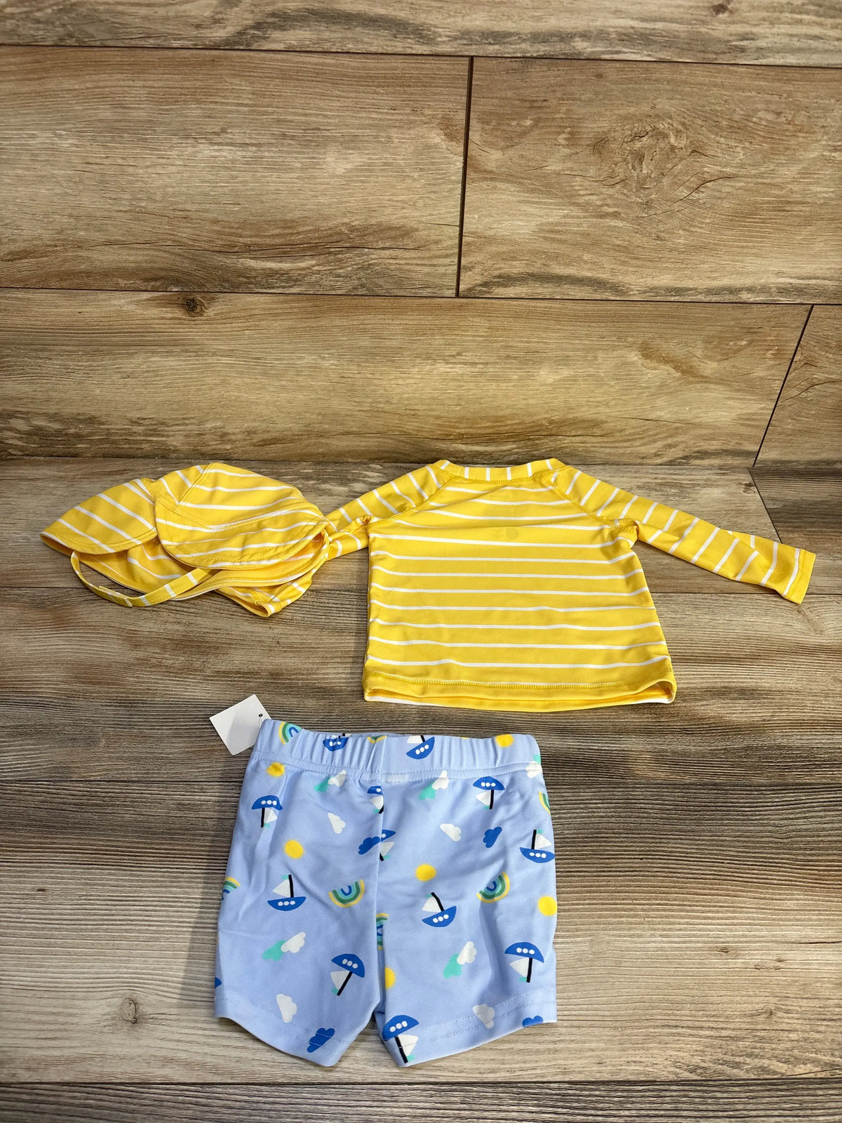 Flat lay of First Impression 3-piece swim set showcasing yellow striped rash guard and hat with patterned blue trunks on wood