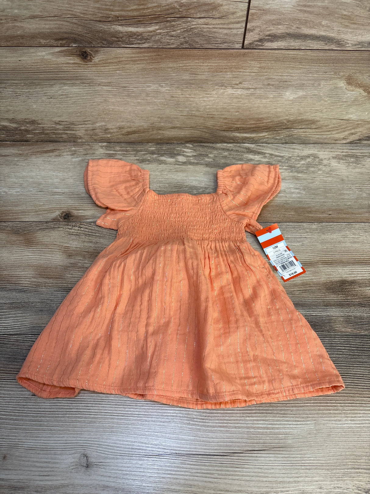Flat lay of peach toddler dress showcasing smocked bodice, flutter sleeves, and gold metallic stripes with wooden background and size 12M tag