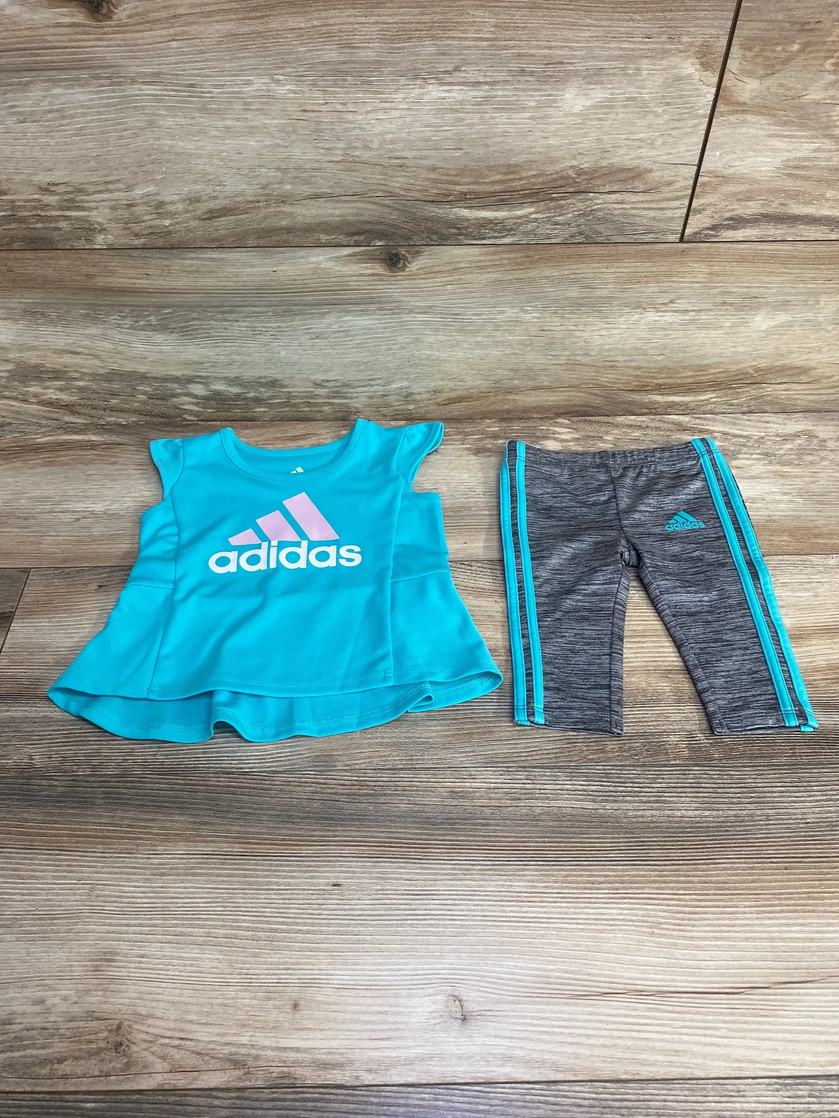 Flat lay of Adidas two-piece: teal flared top with pink Adidas logo and gray leggings with teal side stripes on wood floor