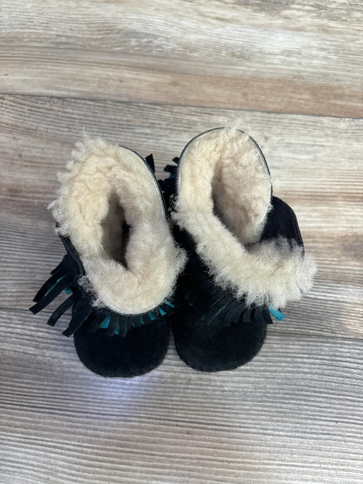 Top view of black baby moccasin boots with cream wool lining and fringe details on wooden floor