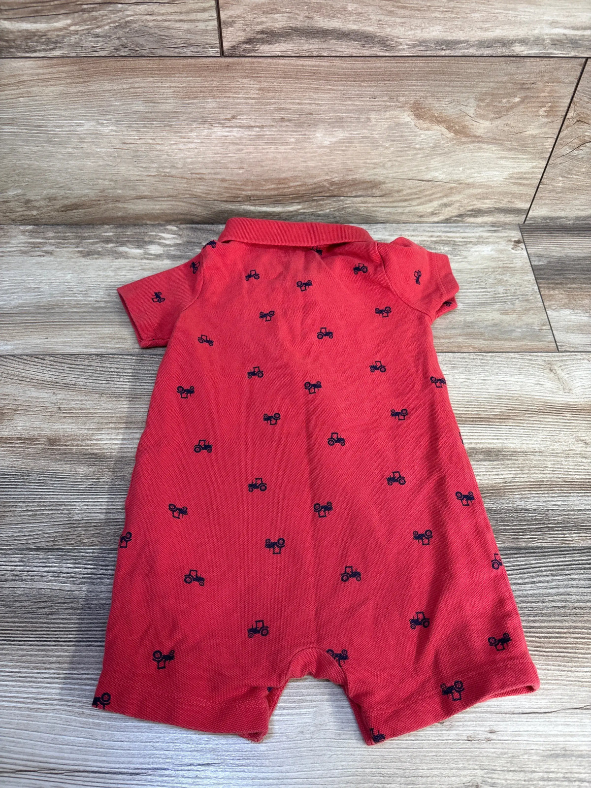 Front view of Carter’s red polo shortie romper showcasing navy tractor pattern, short sleeves, and polo collar on a wood floor