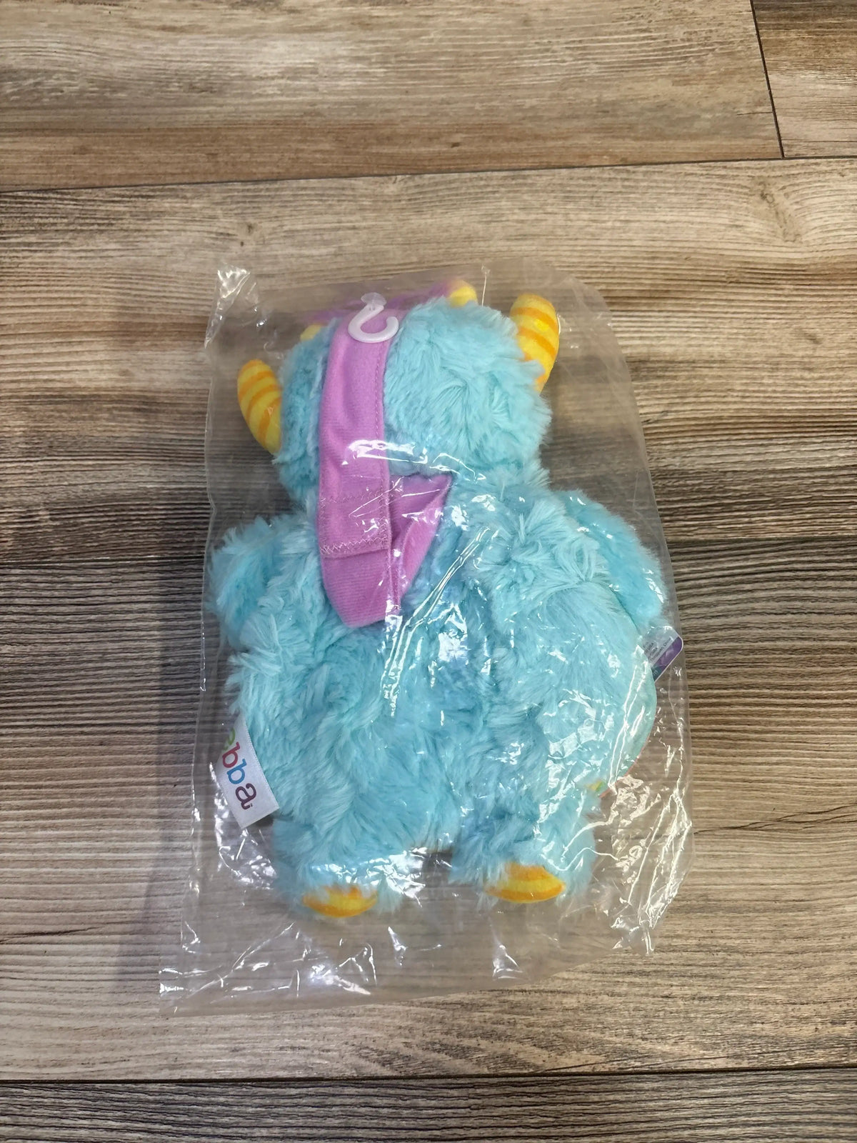 Rear view of ebba Story Pals plush book blue furry monster with yellow horns and purple headband in clear plastic on wood