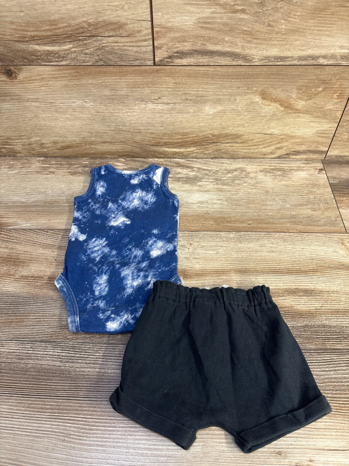 Top view of Carter’s two-piece tank bodysuit and shorts set showcasing a blue tie-dye bodysuit and black shorts on a wood floor