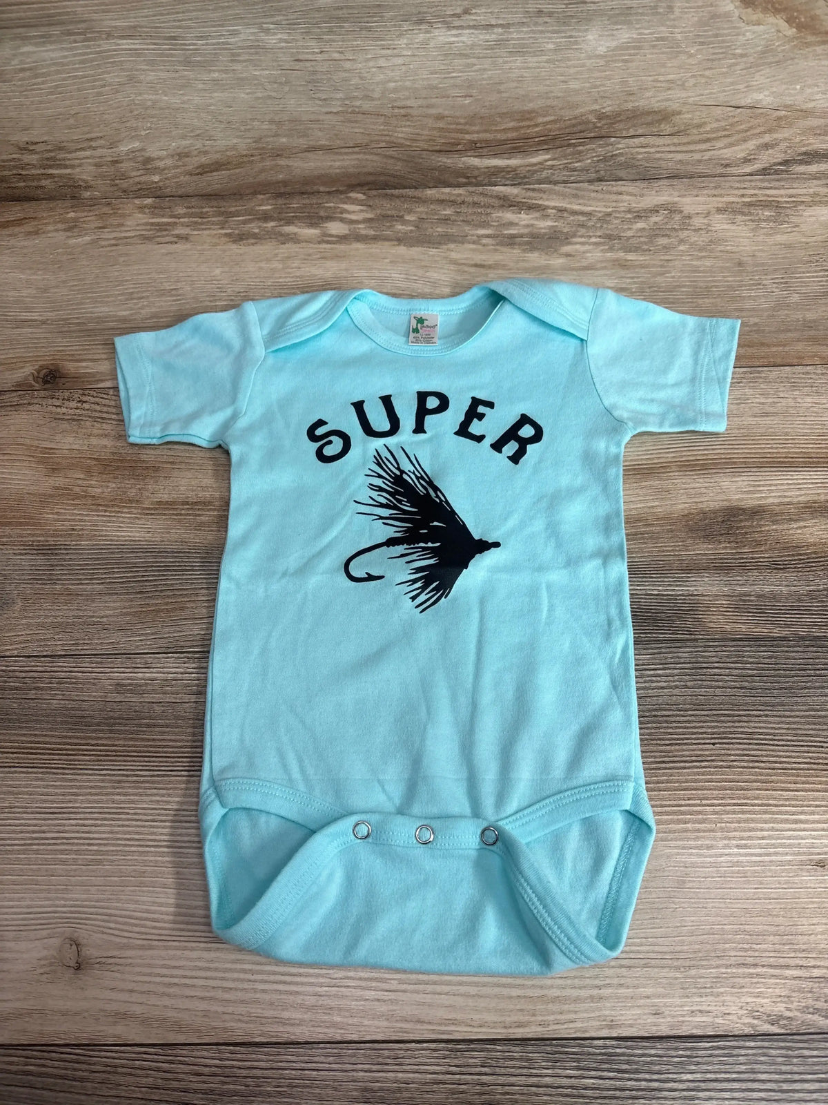 Flat-lay of blue Laughing Giraffe Super Fly bodysuit 12-18 months showing black SUPER text and fishing-fly graphic on wood