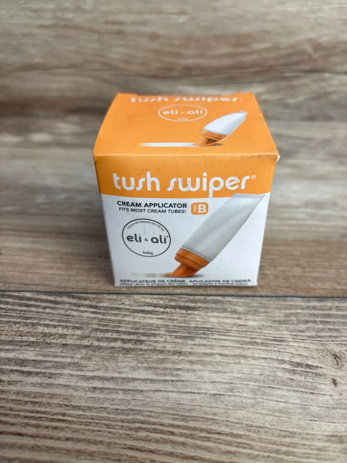 Front view of NEW Eli and Ali Tush Swiper Cream Applicator Type B in original orange and white retail box on wood plank floor