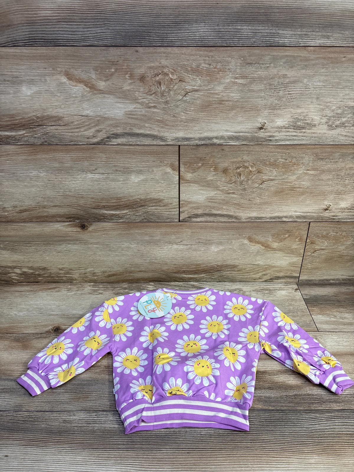 Flat lay of Little Sleepies bamboo daisy pullover sweatshirt showing purple fabric with white daisies and yellow smiling centers on wood background