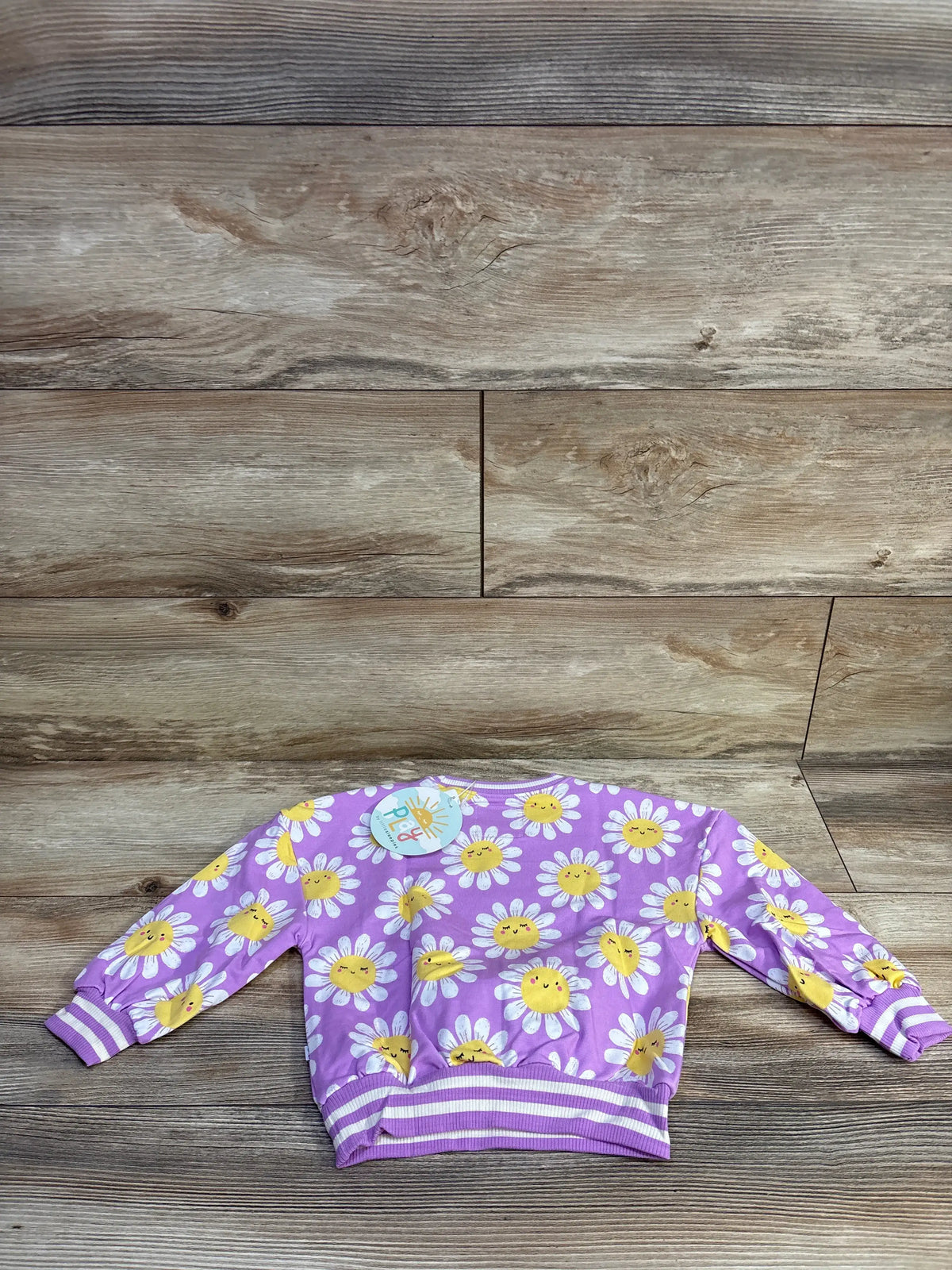 Flat lay of Little Sleepies bamboo daisy pullover sweatshirt showing purple fabric with white daisies and yellow smiling centers on wood background