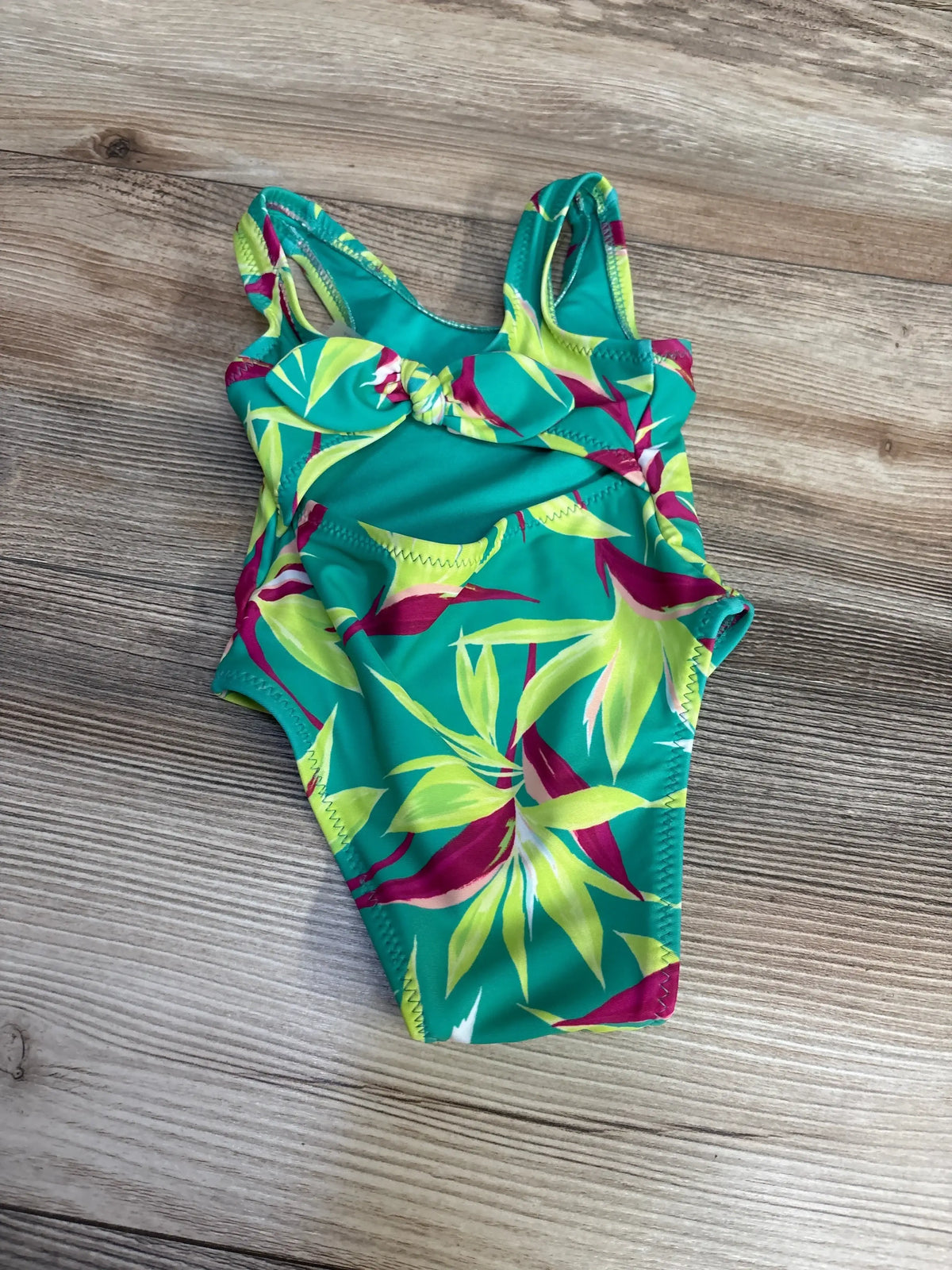 Top-down view of Old Navy newborn one-piece cutout swimsuit, green tropical leaf print, front knot and side cutouts on wood