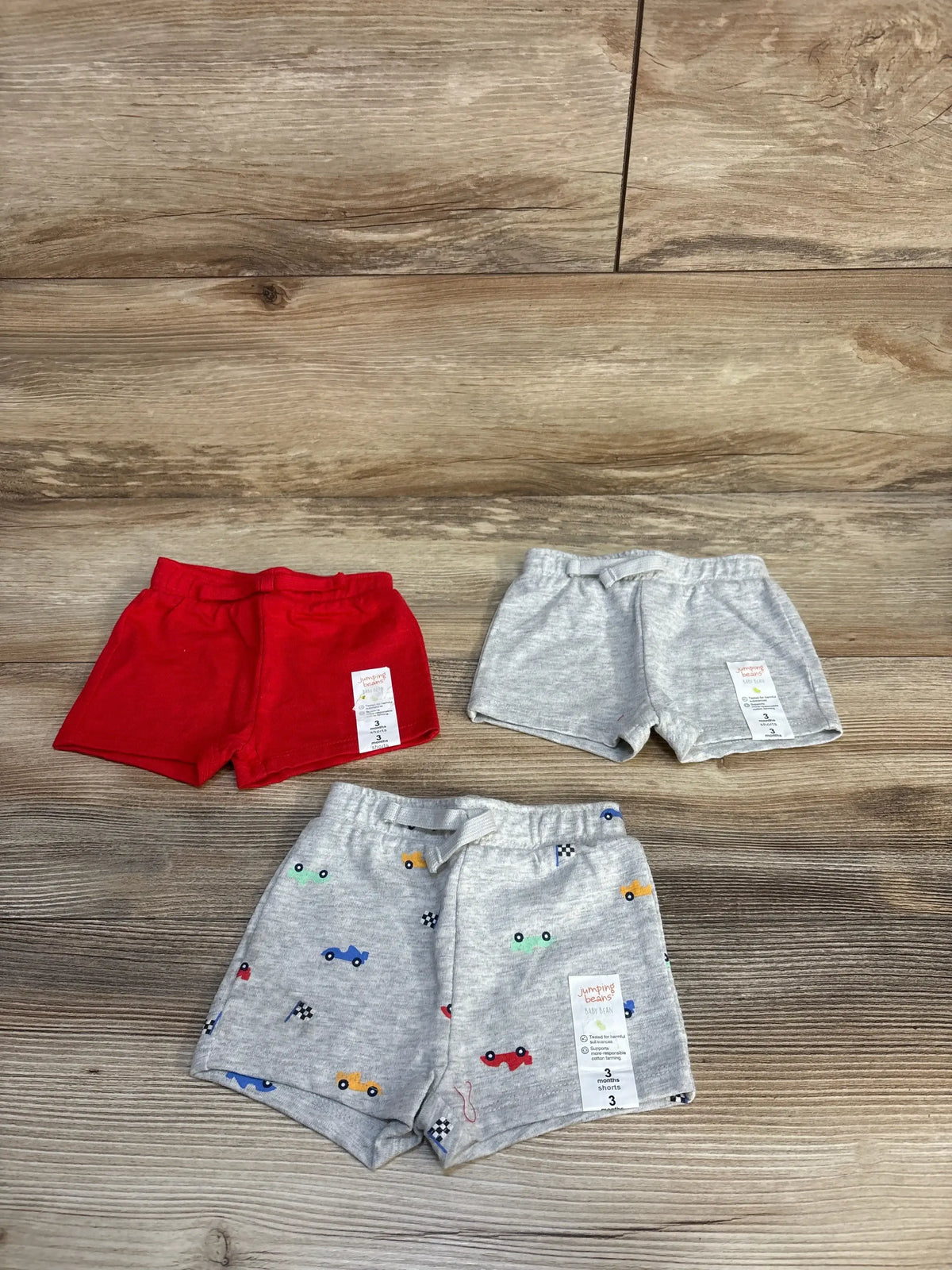 Set of three toddler boys' shorts, red, gray, and gray with colorful cars, on wood floor