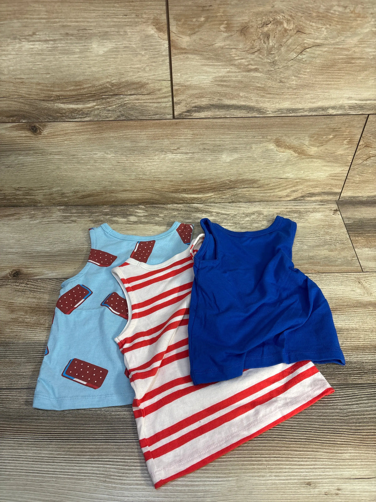 Top-down view of Cat and Jack 3-pack ice cream tank tops showcasing light blue ice cream bar, red-striped, and solid blue designs on a wood floor