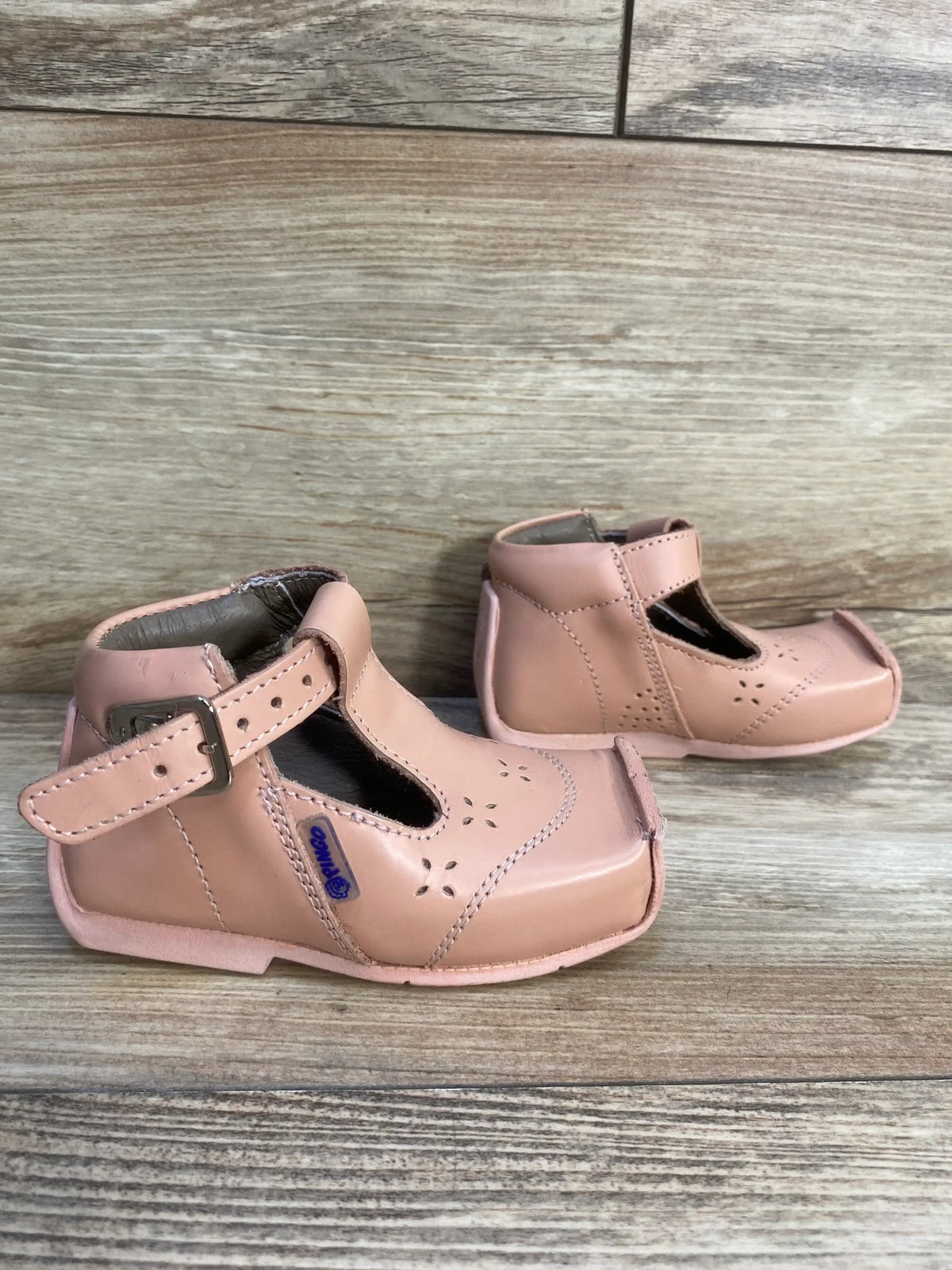 Pink leather toddler shoes with buckle strap and cutout floral accents on wood background