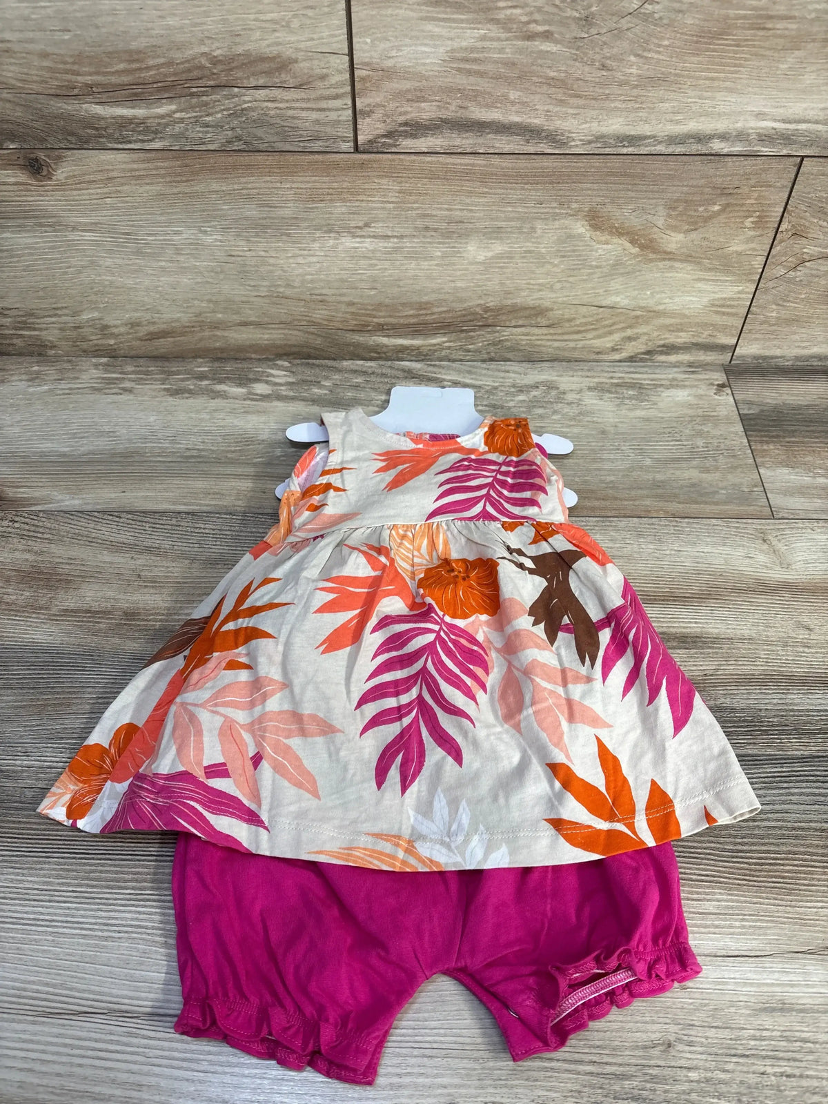 Flat lay of Carter’s 3-piece baby dress set showcasing a beige sleeveless tropical leaf dress with gathered skirt and magenta ruffle bloomers on light wood floor