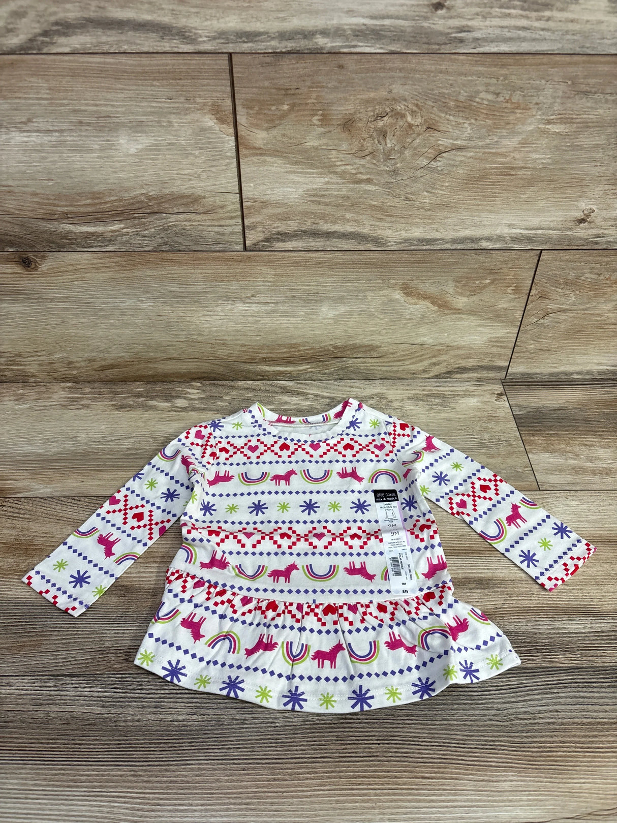 Flat lay of white peplum baby shirt with pink unicorns, rainbows, and fair-isle motifs, 9M tag on wood floor