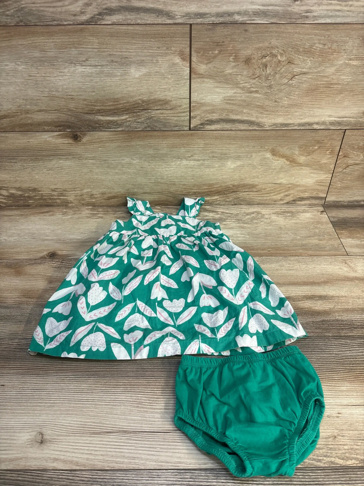 Flat lay of green baby dress with matching bloomers, white floral print and ruffle shoulder straps on wood background