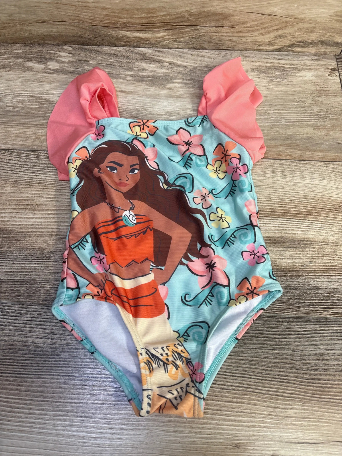Front view of toddler one-piece Moana swimsuit with large Moana print, aqua floral background and coral ruffle sleeves on wood