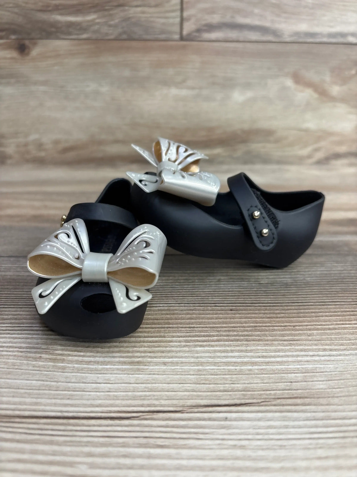Angled view of Mini Melissa Ultragirl Bow black jelly Mary Jane flats size 5C showcasing gray decorative bows on wood background