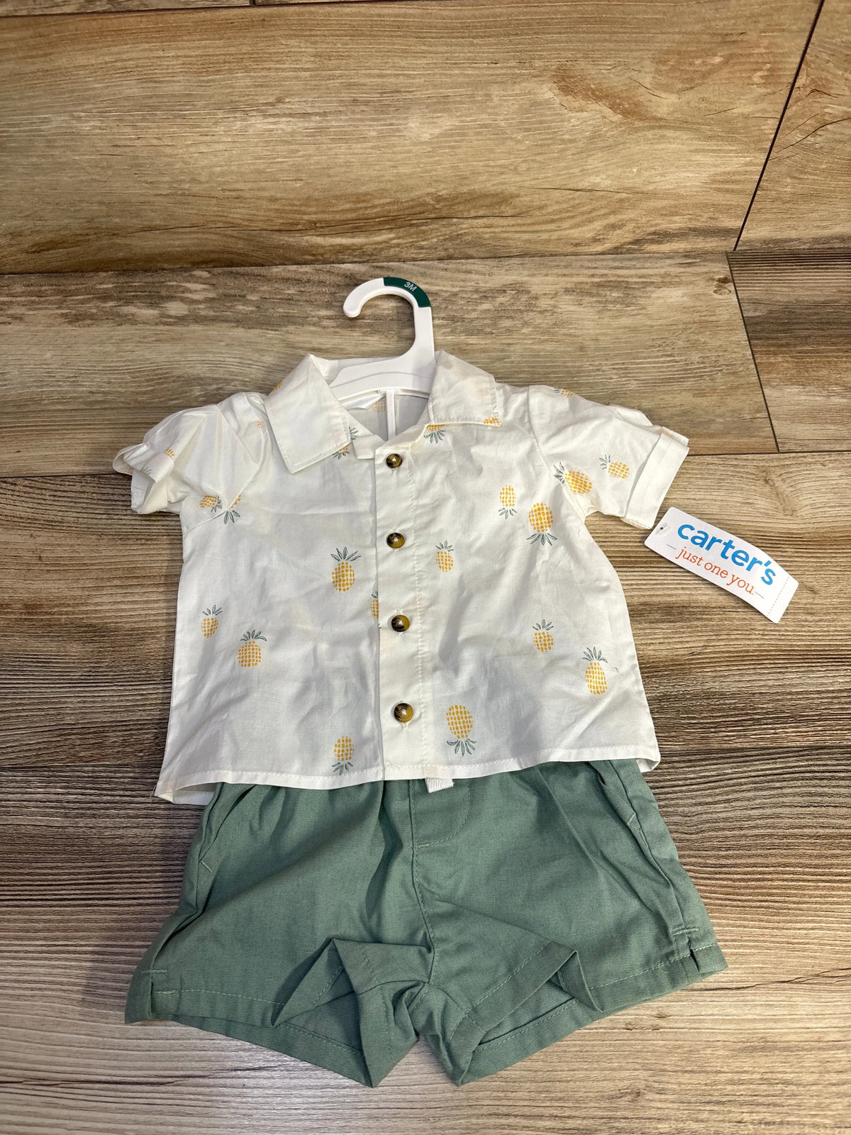 Flat lay of infant pineapple-print button-up shirt and sage green shorts showcasing front buttons and collar on wood background
