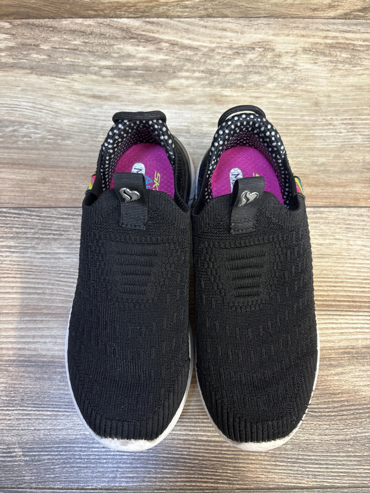 Black slip-on kids sneakers with white soles and pink insoles on wooden floor