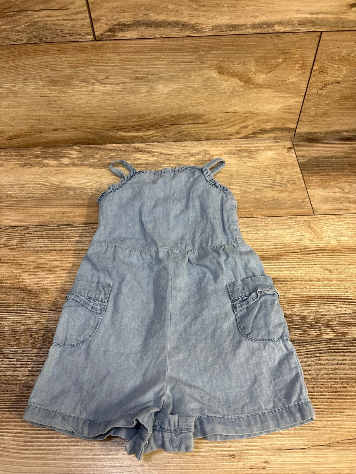 Flat-lay of blue chambray toddler romper showing ruffle shoulder straps, front patch pockets, and snap crotch on wood floor